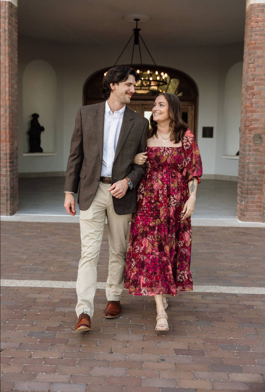 Couple walking arm in arm, woman in floral red dress, man in brown blazer, brick courtyard backdrop.