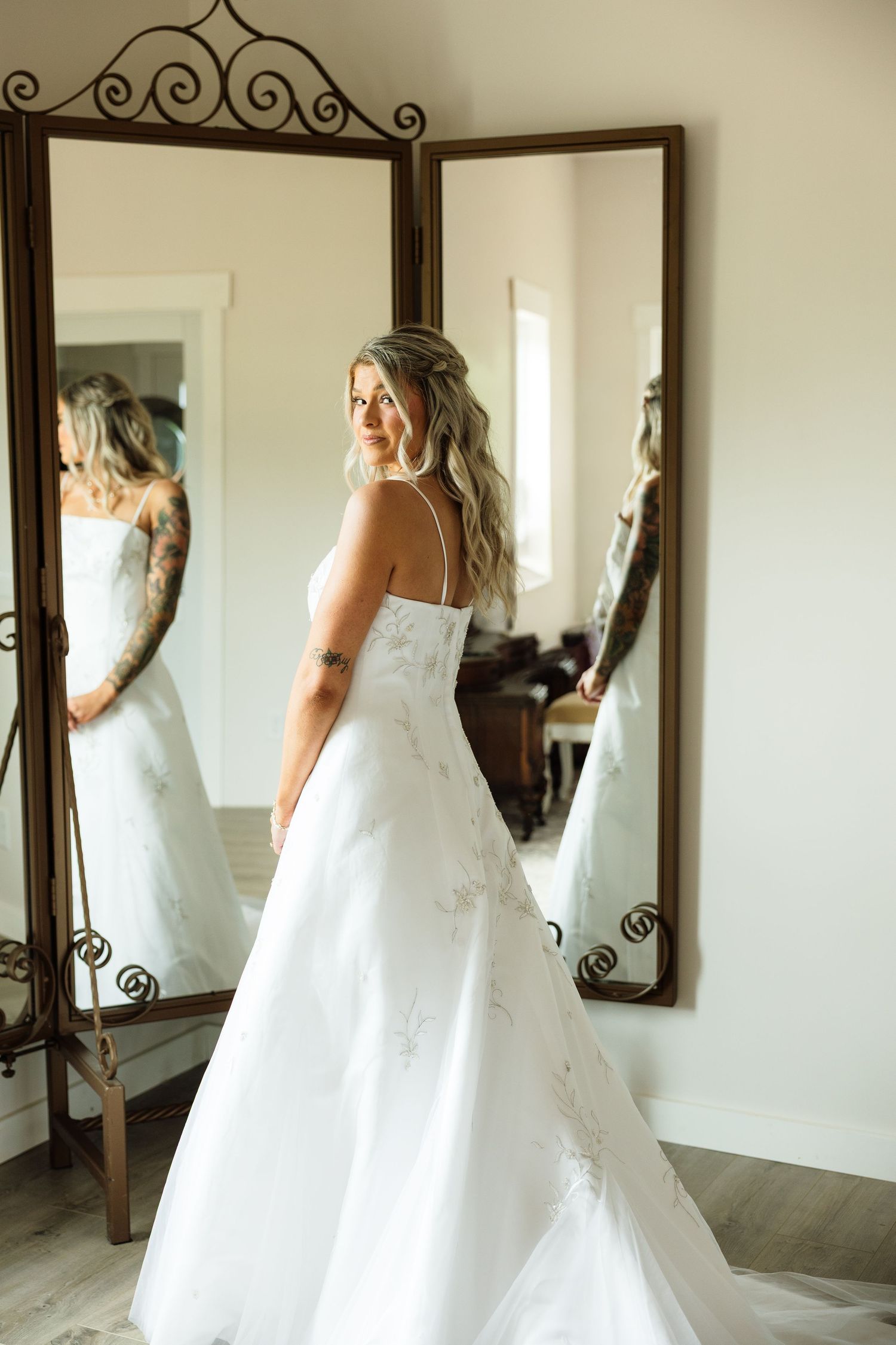 Bride in a strapless white A-line wedding gown standing before a tri-fold mirror, smiling over her shoulder.