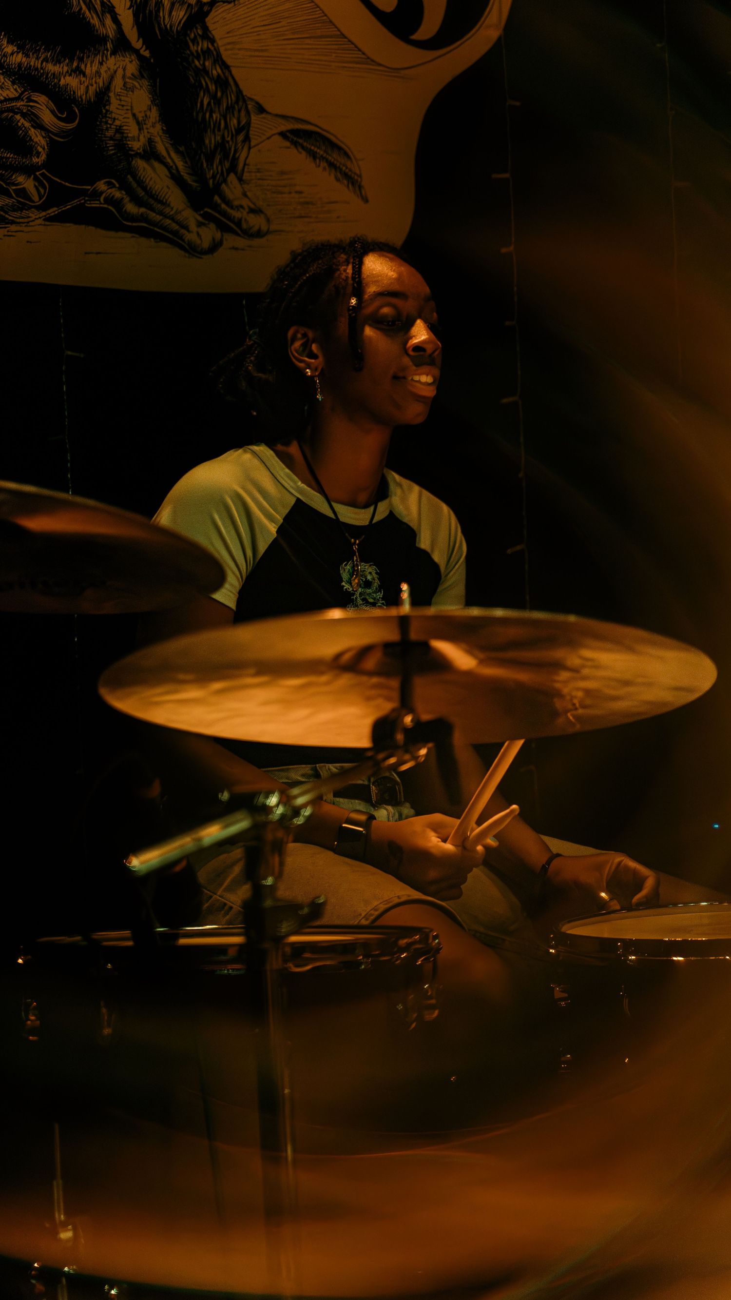 A drummer performs energetically behind a drum kit in warm lighting during a live performance.