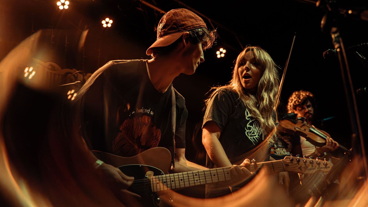 Musicians perform on stage with guitars and instruments in a dimly lit concert venue.