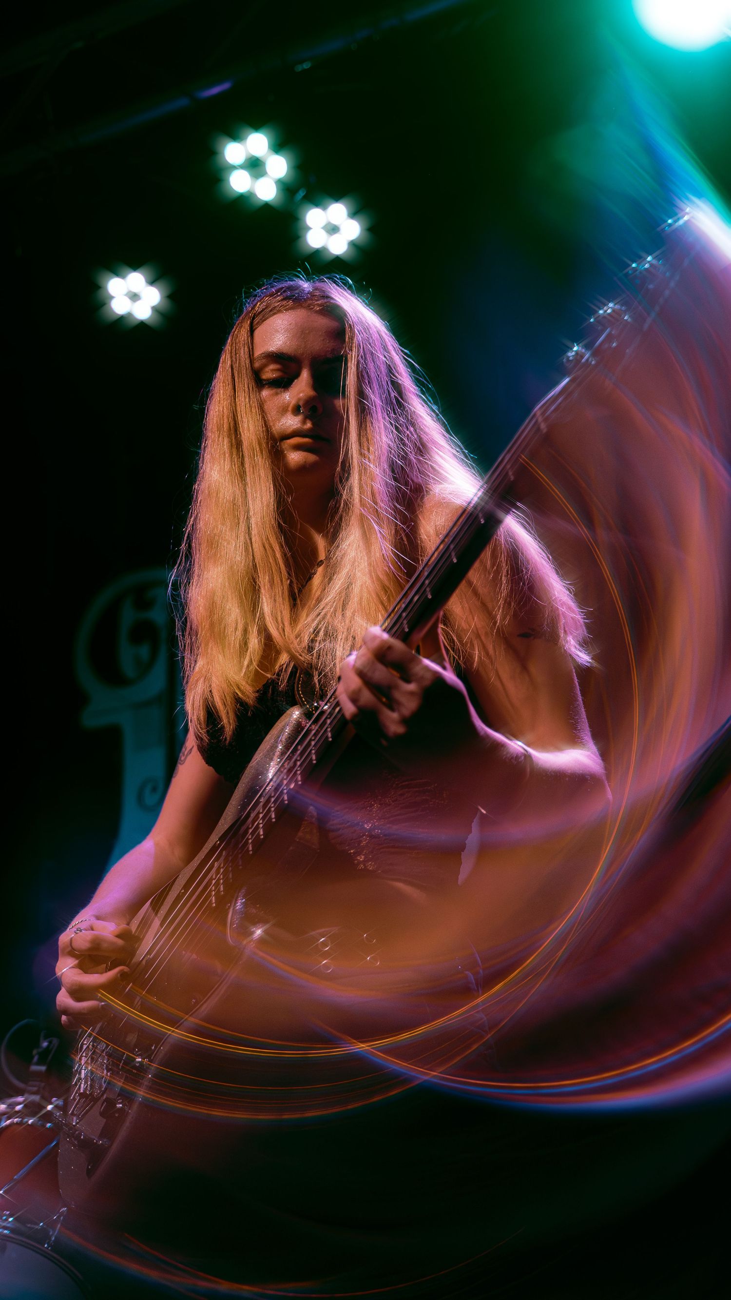 A bass guitarist performs on stage with long blonde hair and dramatic lighting creating motion blur effects in purple and teal.