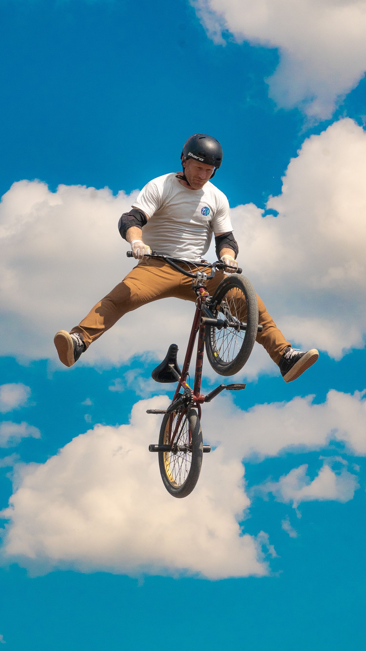 Rider performs no-handed BMX jump trick with legs spread against blue sky with clouds.