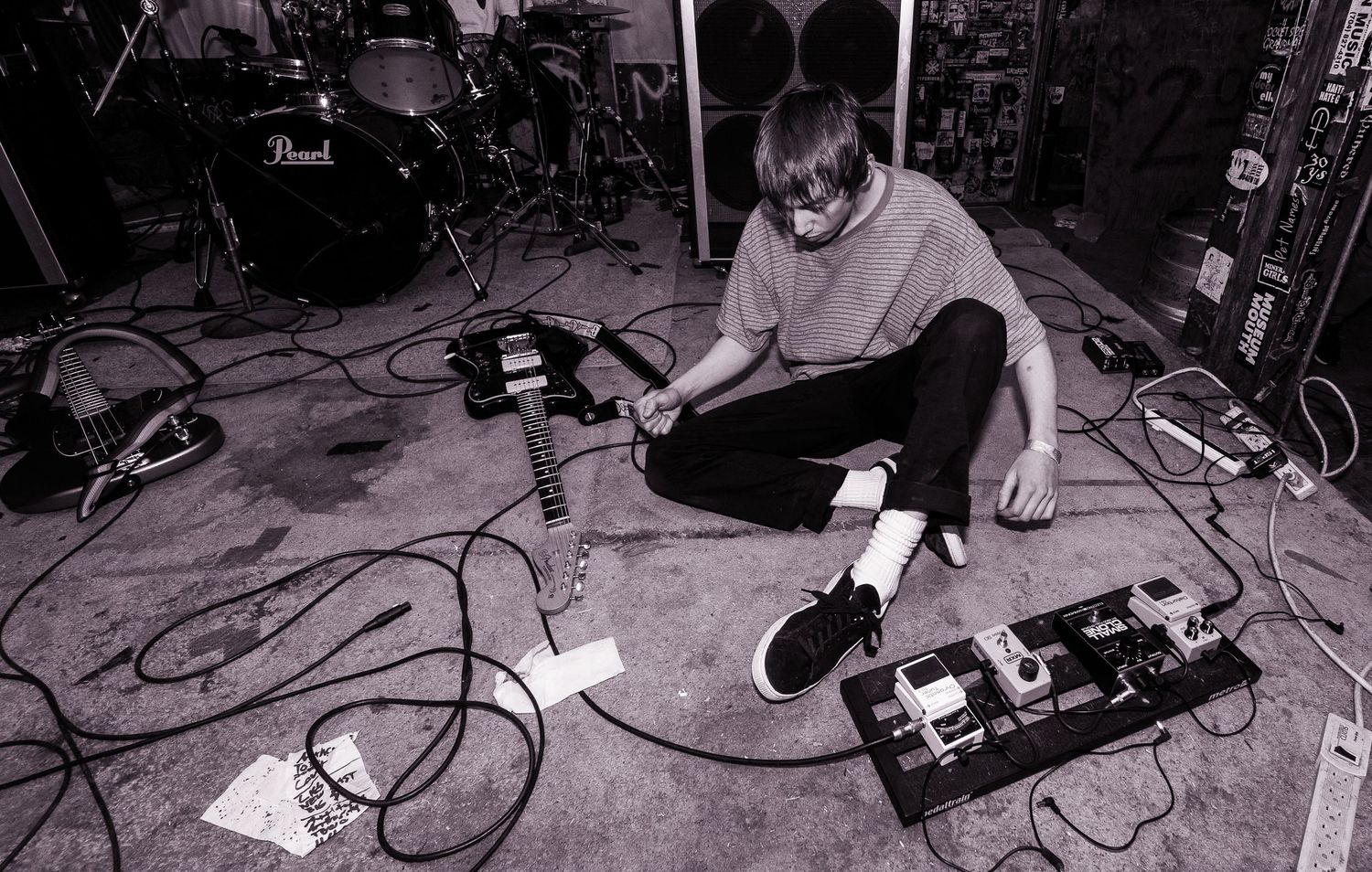 Black and white photo of a musician sitting on the floor adjusting equipment during a performance setup.