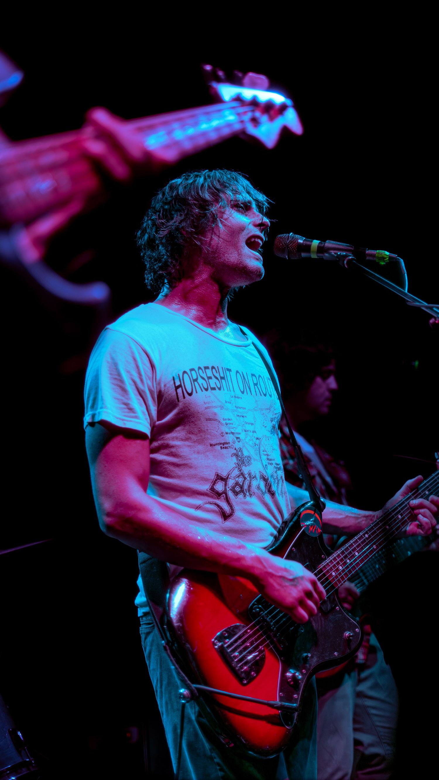 Musicians perform live on stage with guitars under vibrant red and blue lighting effects during a concert.