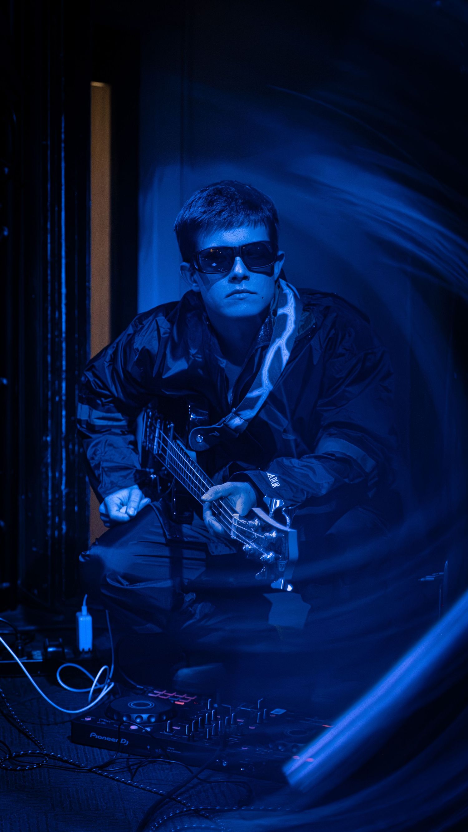 A musician performs with synthesizer equipment under dramatic blue lighting while wearing sunglasses and dark clothing.