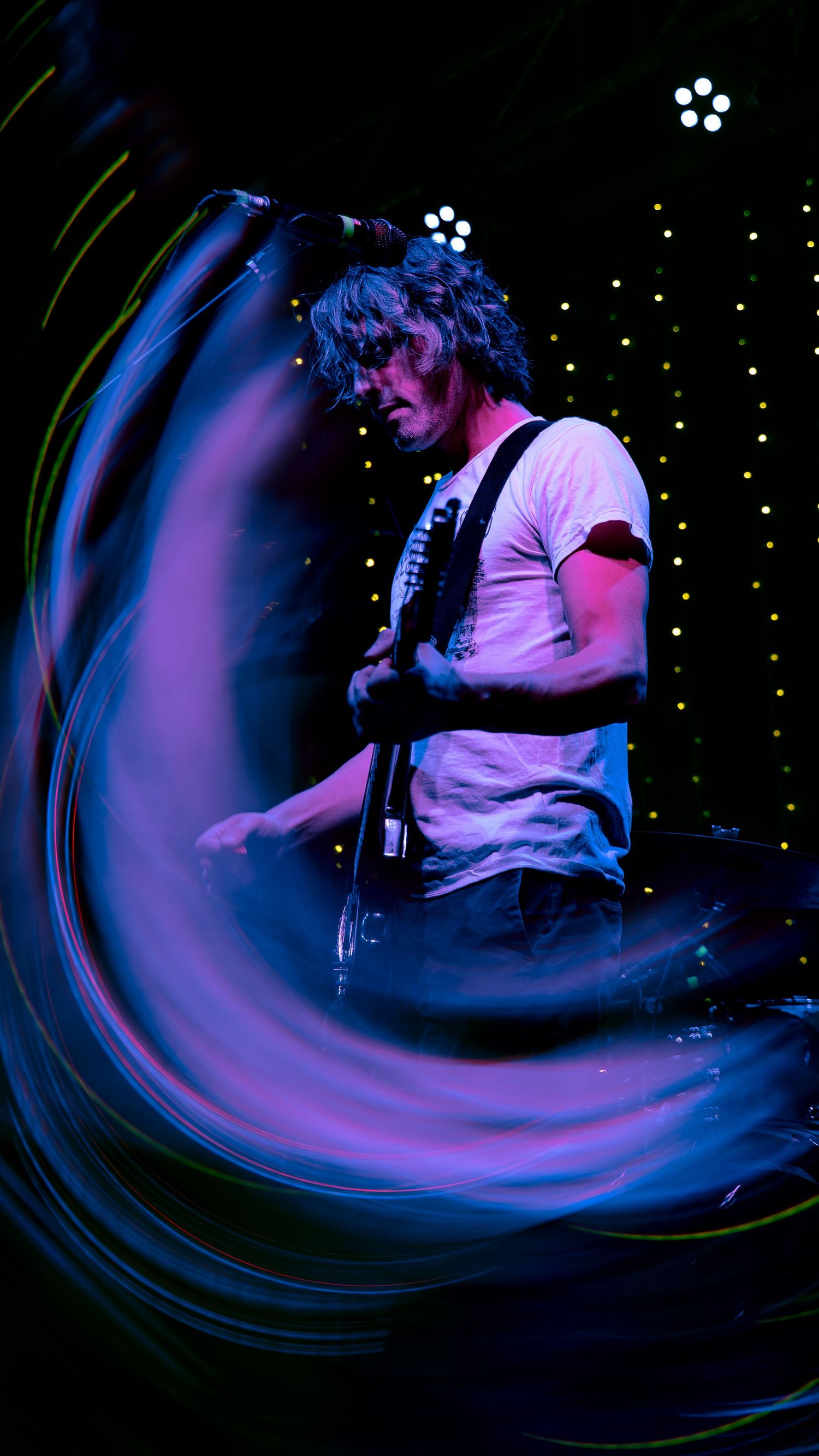 A musician in a white shirt performs on stage with swirling blue and purple lighting effects creating a dynamic visual motion.