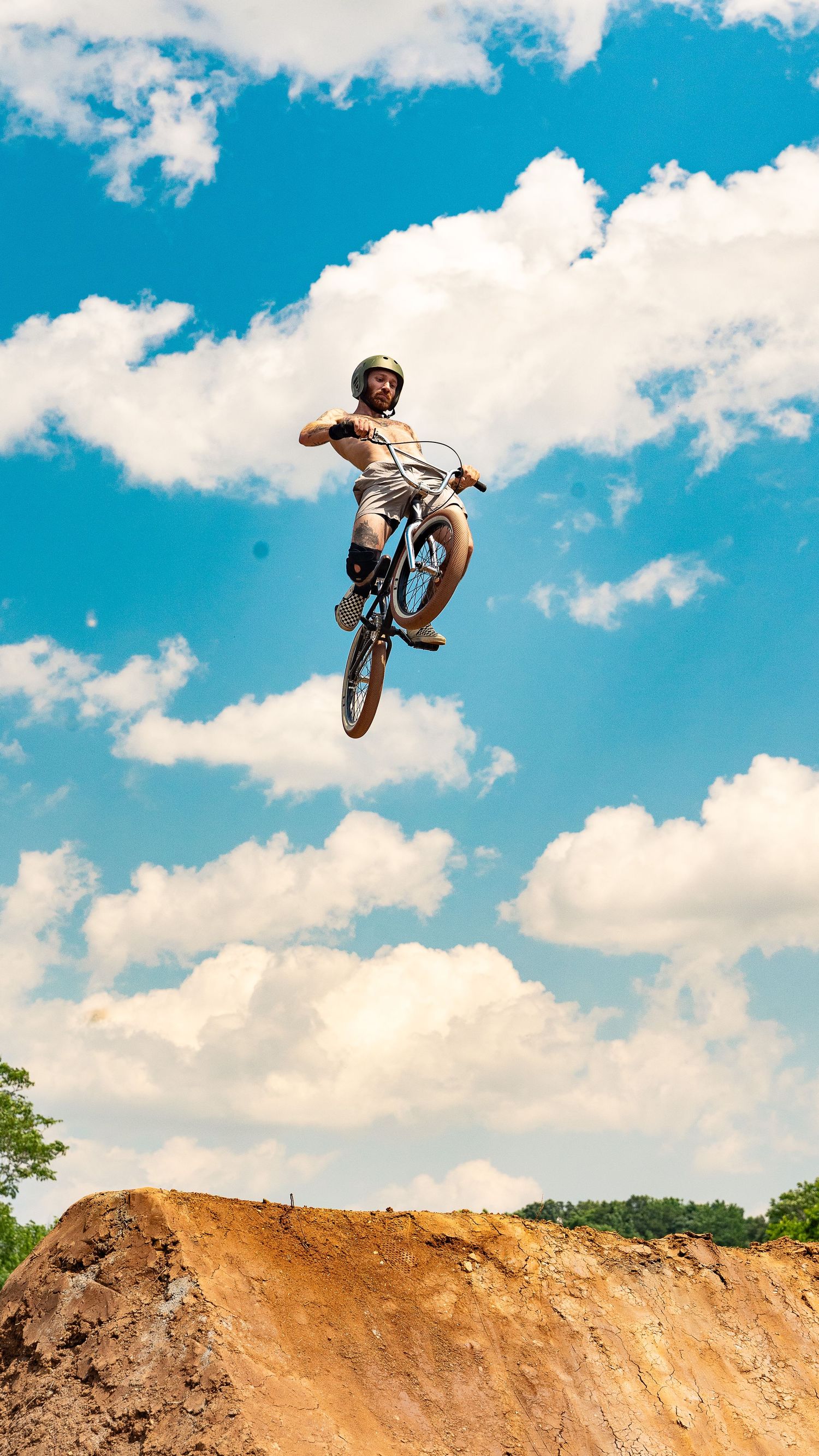 BMX rider launches high above dirt jump performing aerial trick against blue sky with clouds.