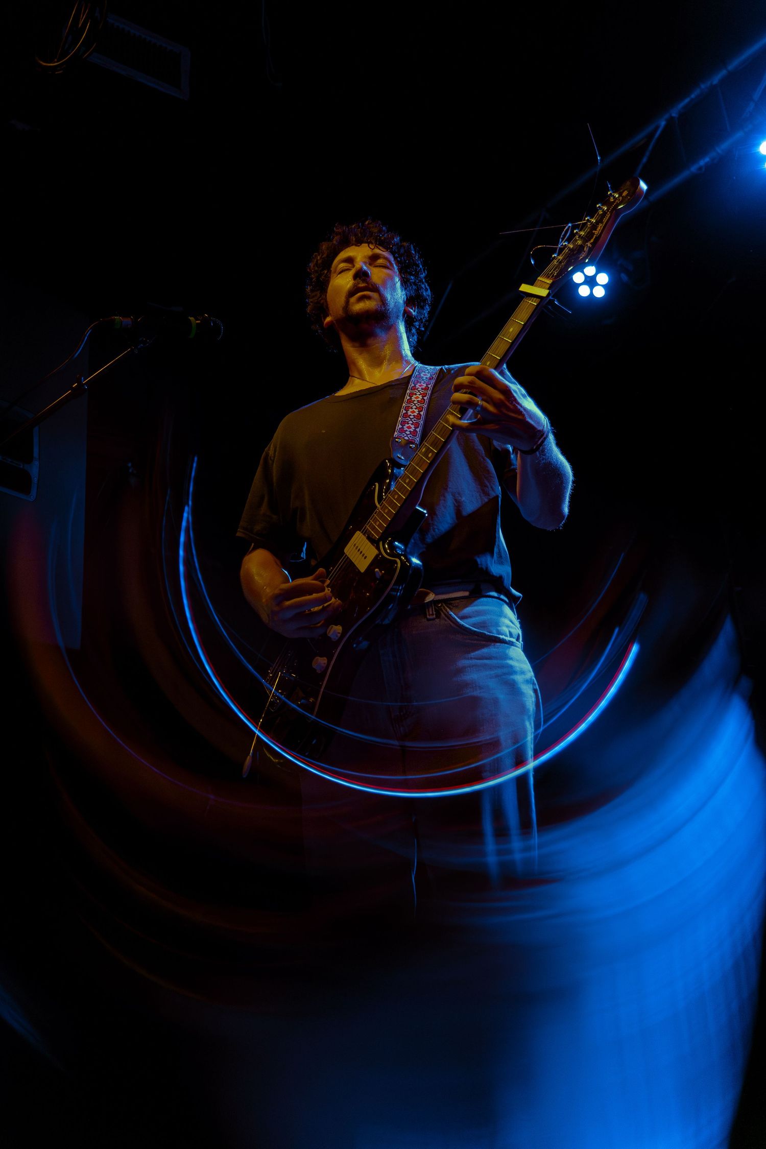 A guitarist plays in dramatic blue stage lighting with swirling motion trails creating an ethereal performance atmosphere.
