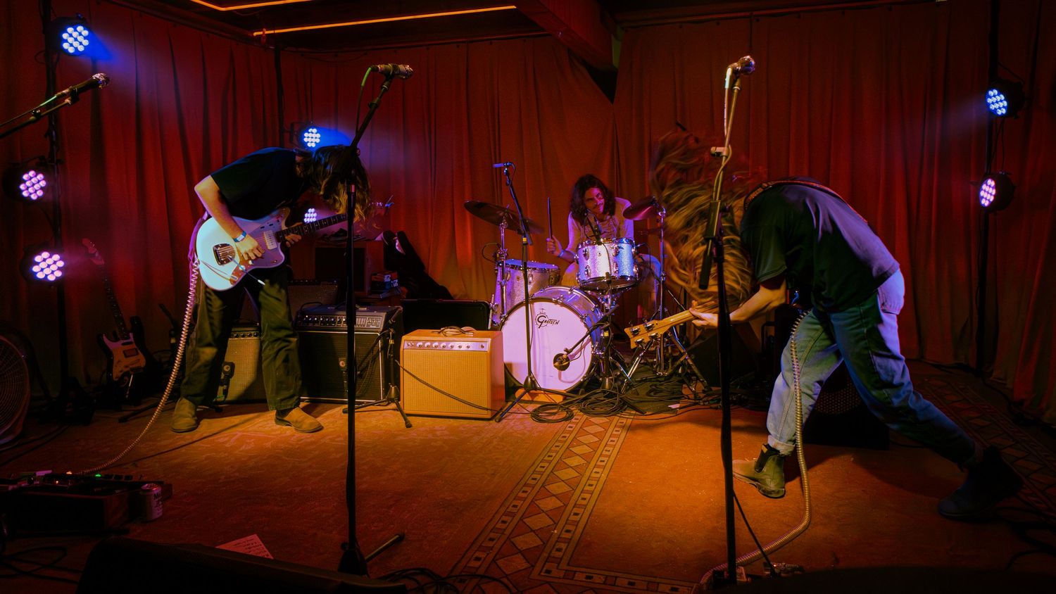 Musicians perform in a red-lit theater illuminated by multiple blue stage lights and spotlights during a live performance.