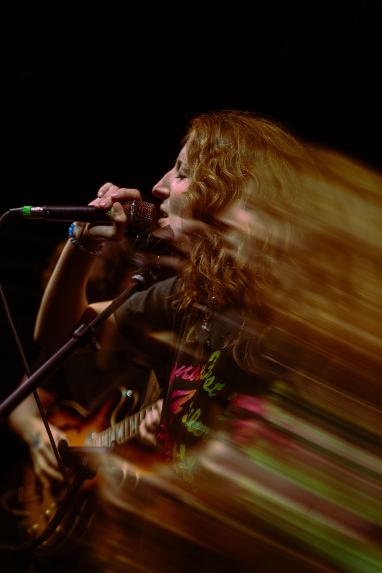 Dynamic concert photo showing a performer with long hair in motion singing into a microphone against a dark background.