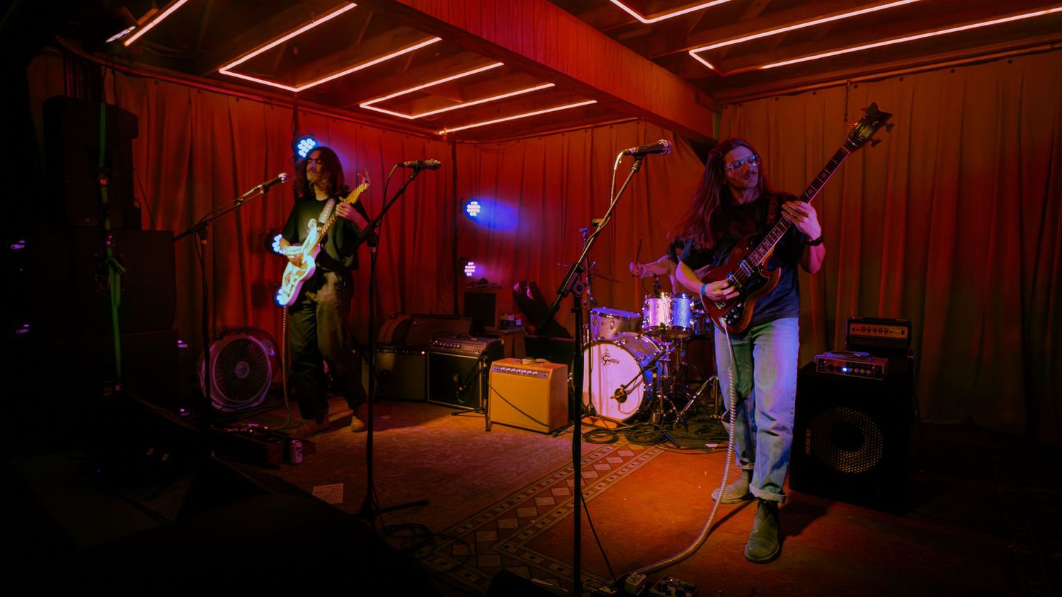 Musicians perform on stage in a red-lit concert venue with electric instruments and microphones setup visible.