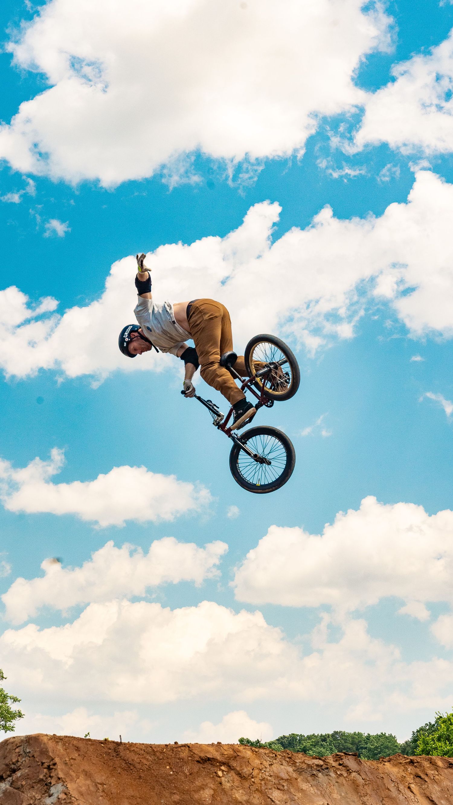 BMX rider performs backflip trick over dirt jump with cloudy sky background.