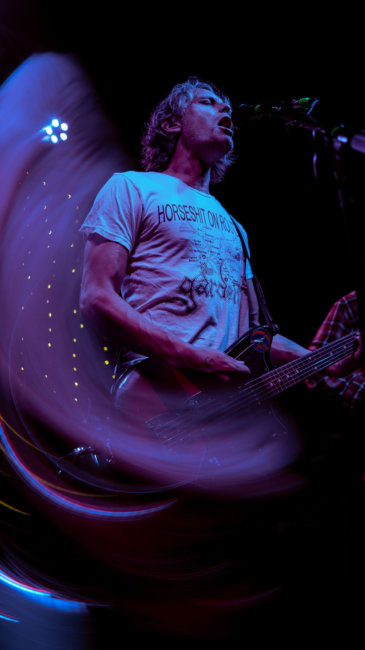Musician in white t-shirt performs energetically on stage with blue lighting effects and bass guitar.