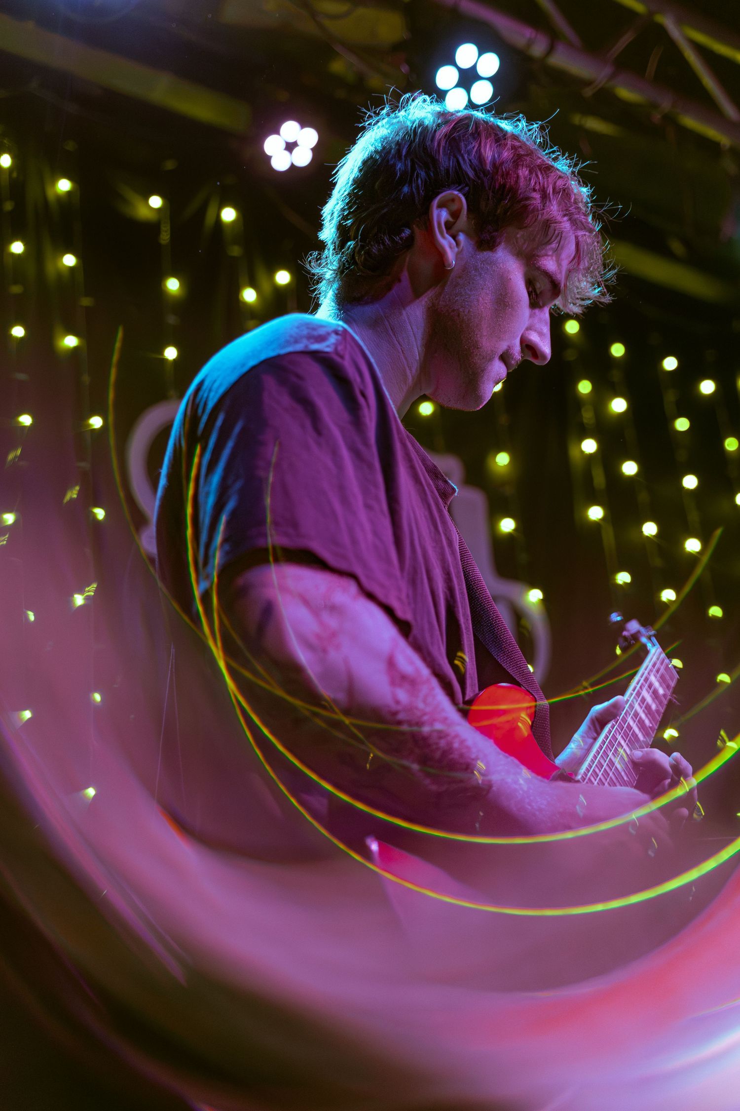 A musician plays guitar on stage under purple lighting with a curved motion blur effect and twinkling lights in background.