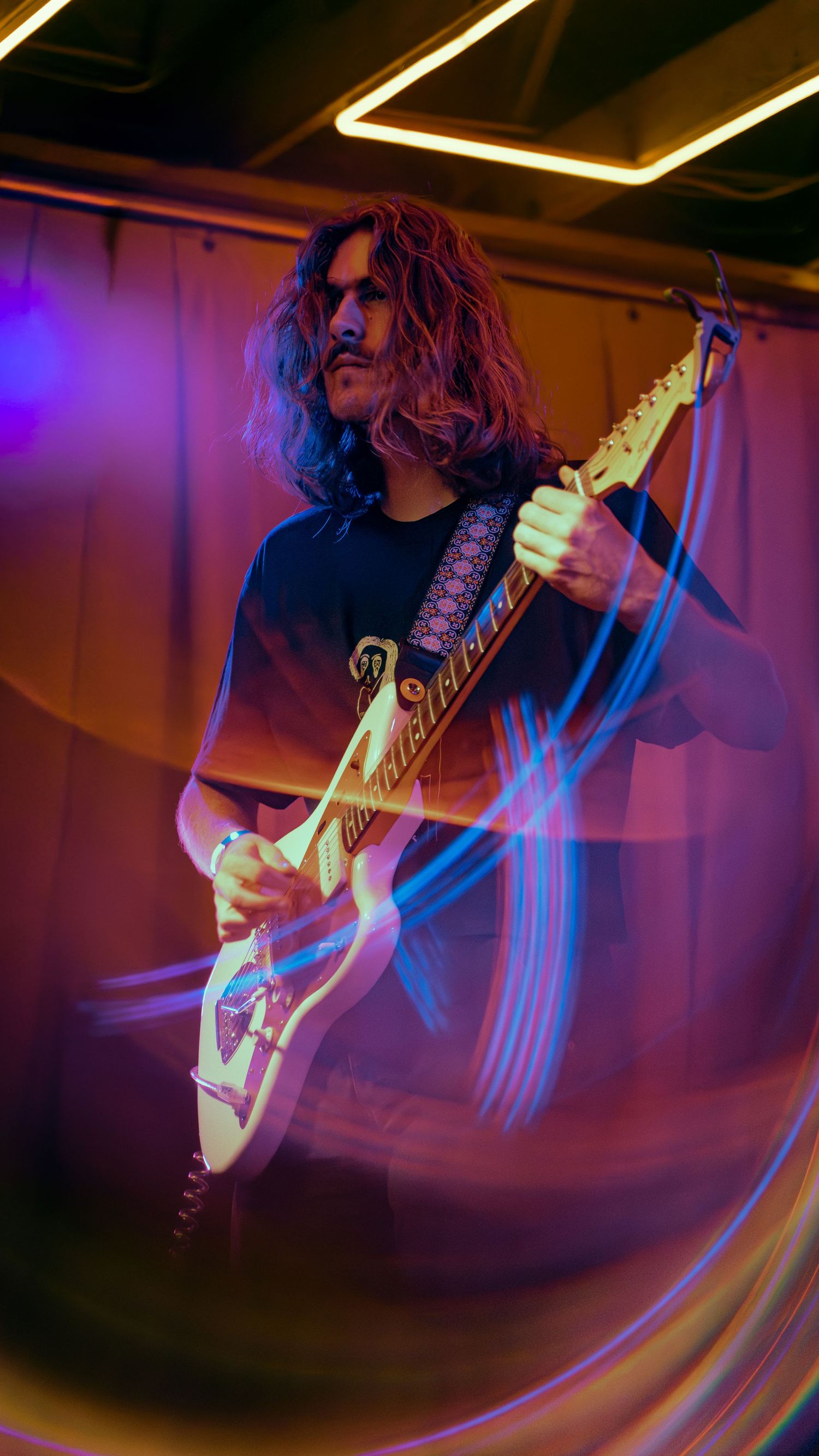 Dynamic performance photo showing guitarist playing electric guitar with colorful purple and blue light trails in motion.
