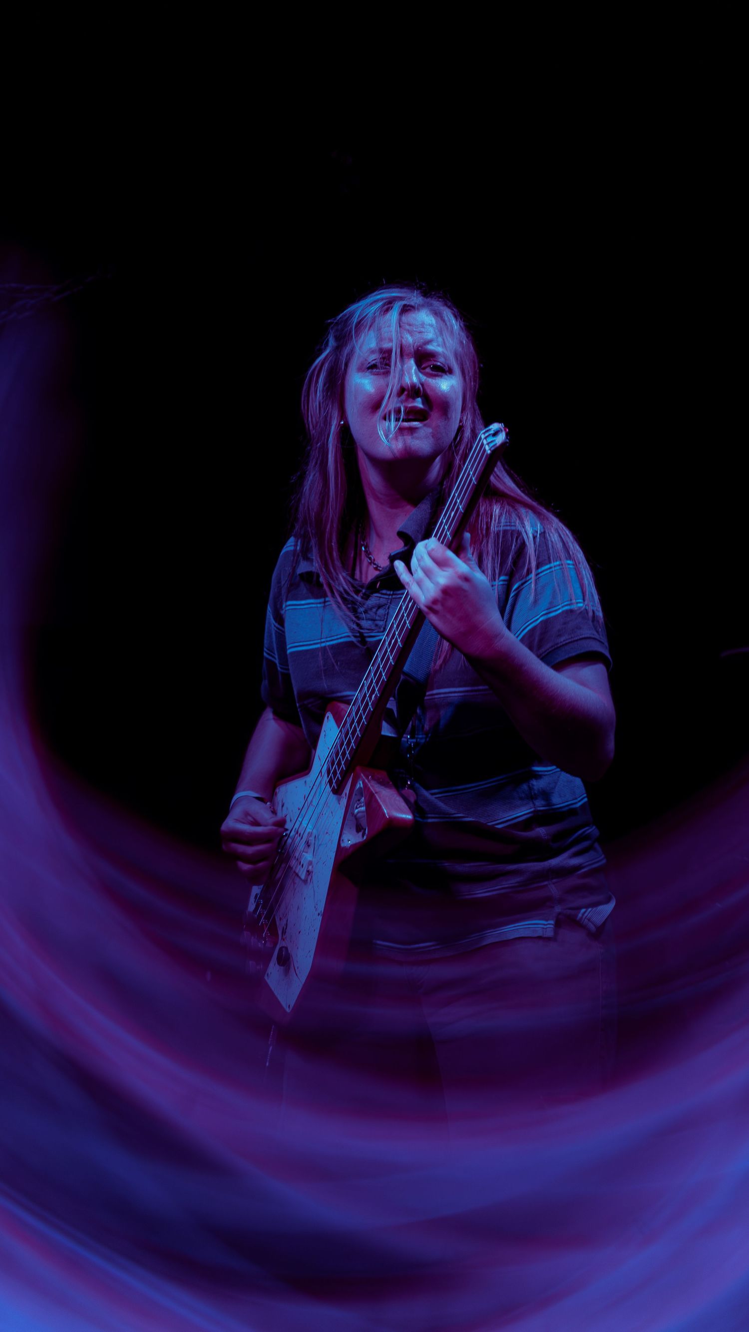 Artist performs on stage with a guitar bathed in dramatic purple and blue concert lighting.