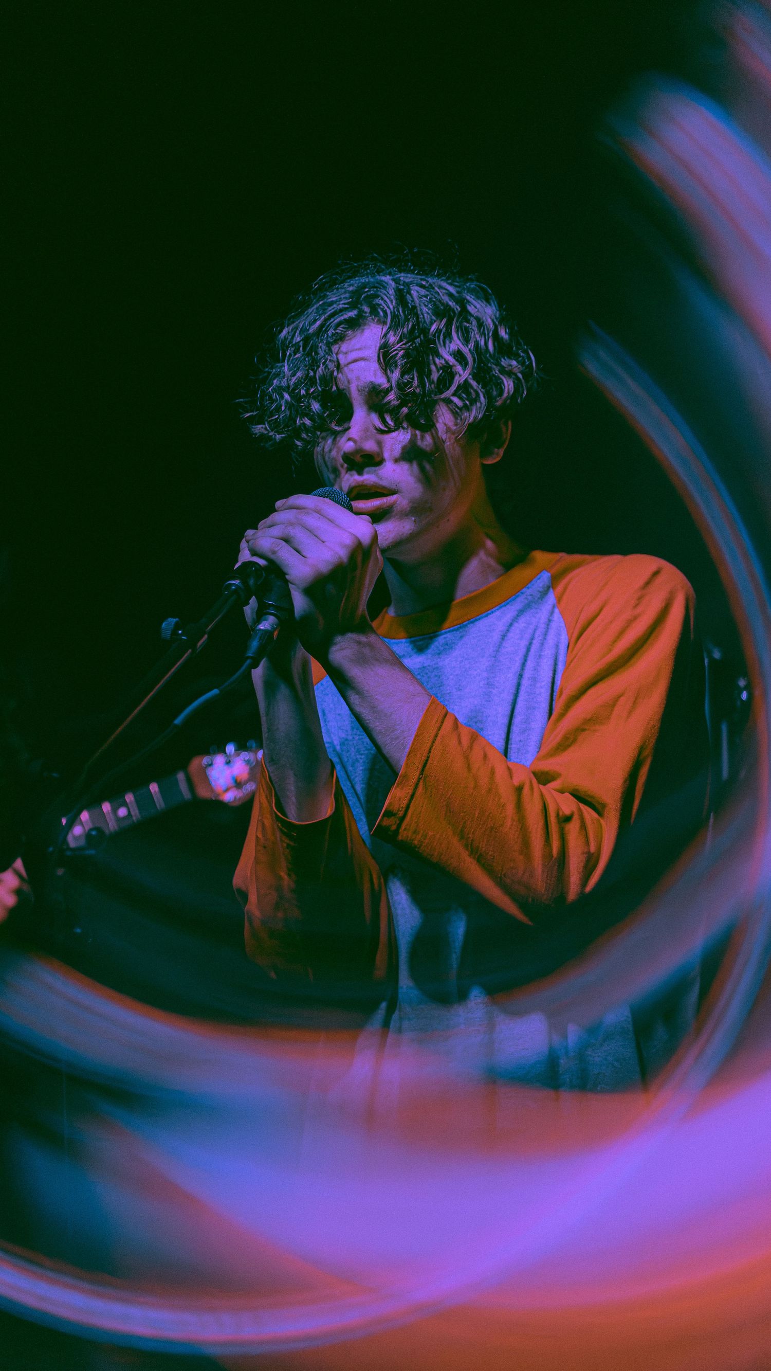 A performer singing into a microphone in dramatic purple and orange lighting during a moody concert performance.