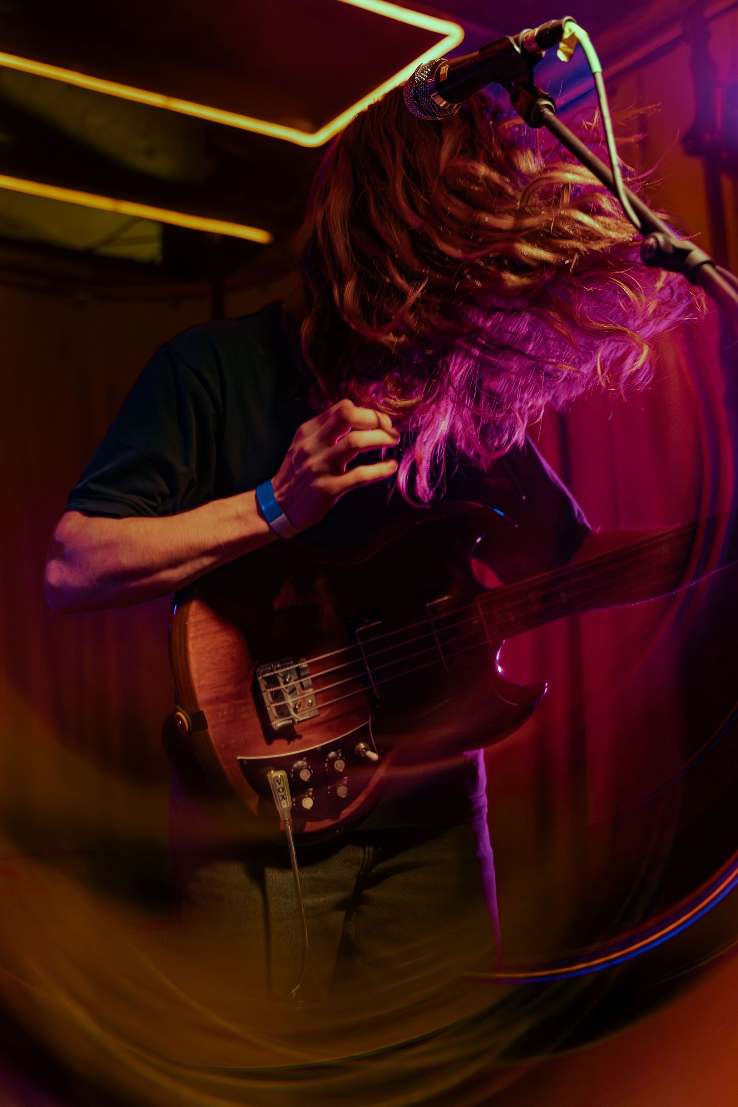 Bass guitarist performing on stage with flowing hair bathed in vibrant purple and yellow stage lighting.