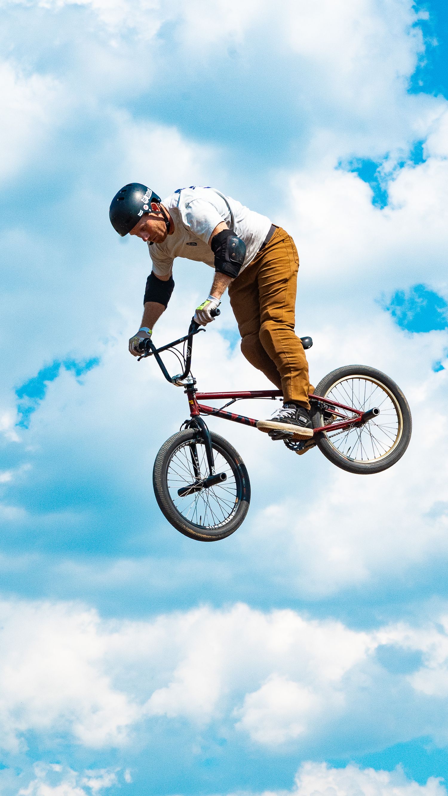 BMX rider soars through the air performing a stylish jump trick with puffy clouds in background.