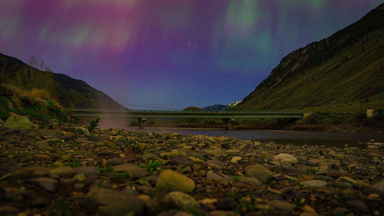 Northern lights dance across the night sky above a rocky beach between mountain peaks.