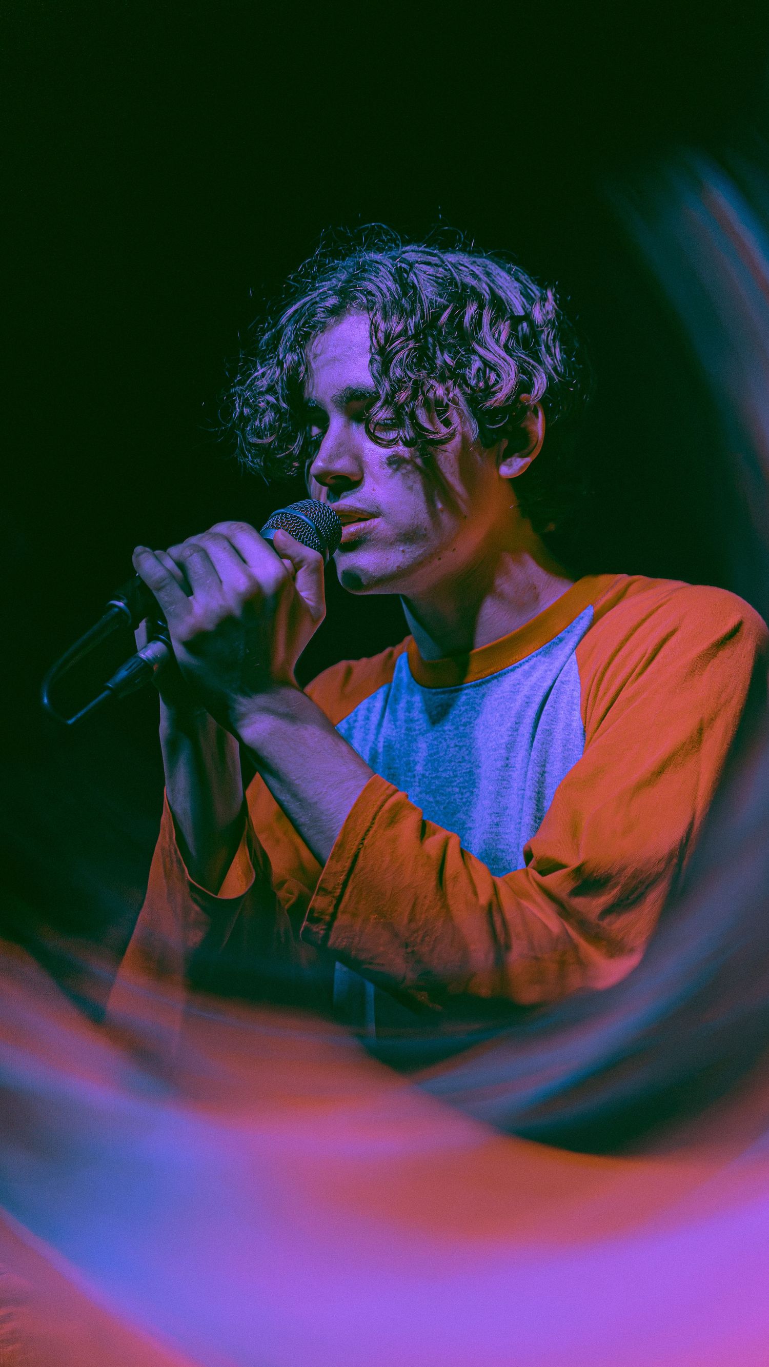 Singer performs on stage in orange jacket under dramatic purple and blue lighting during concert performance.