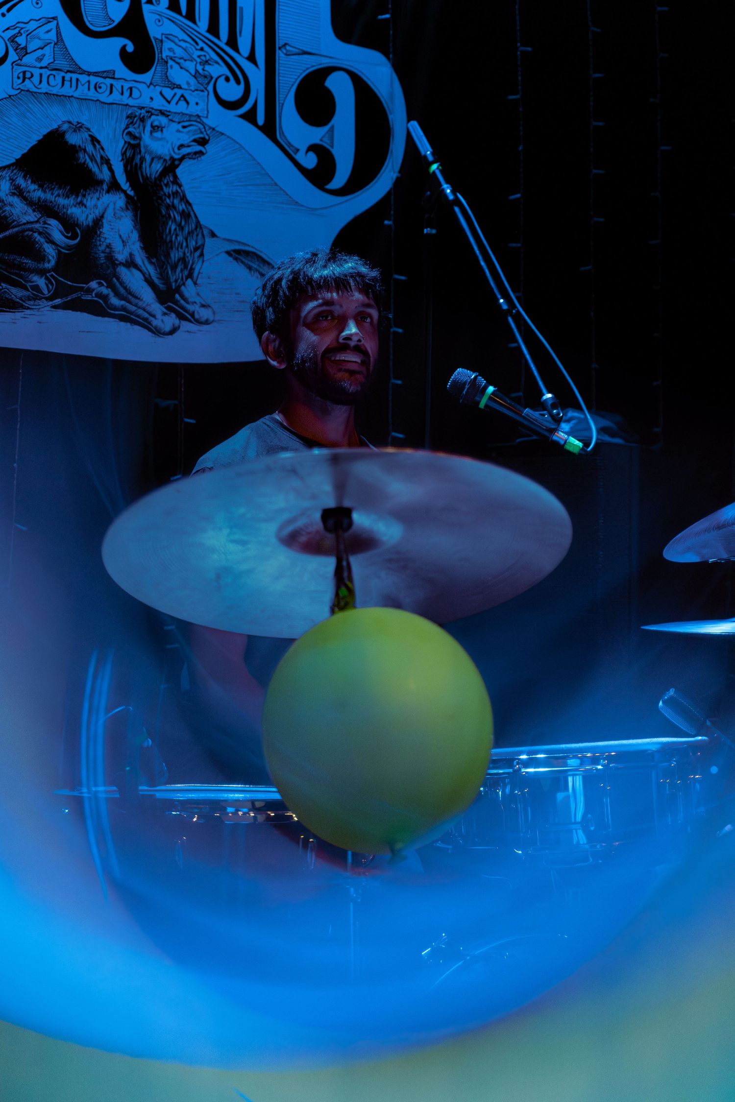 A drummer performs on stage illuminated by dramatic blue lighting with a yellow balloon visible near the drum kit.