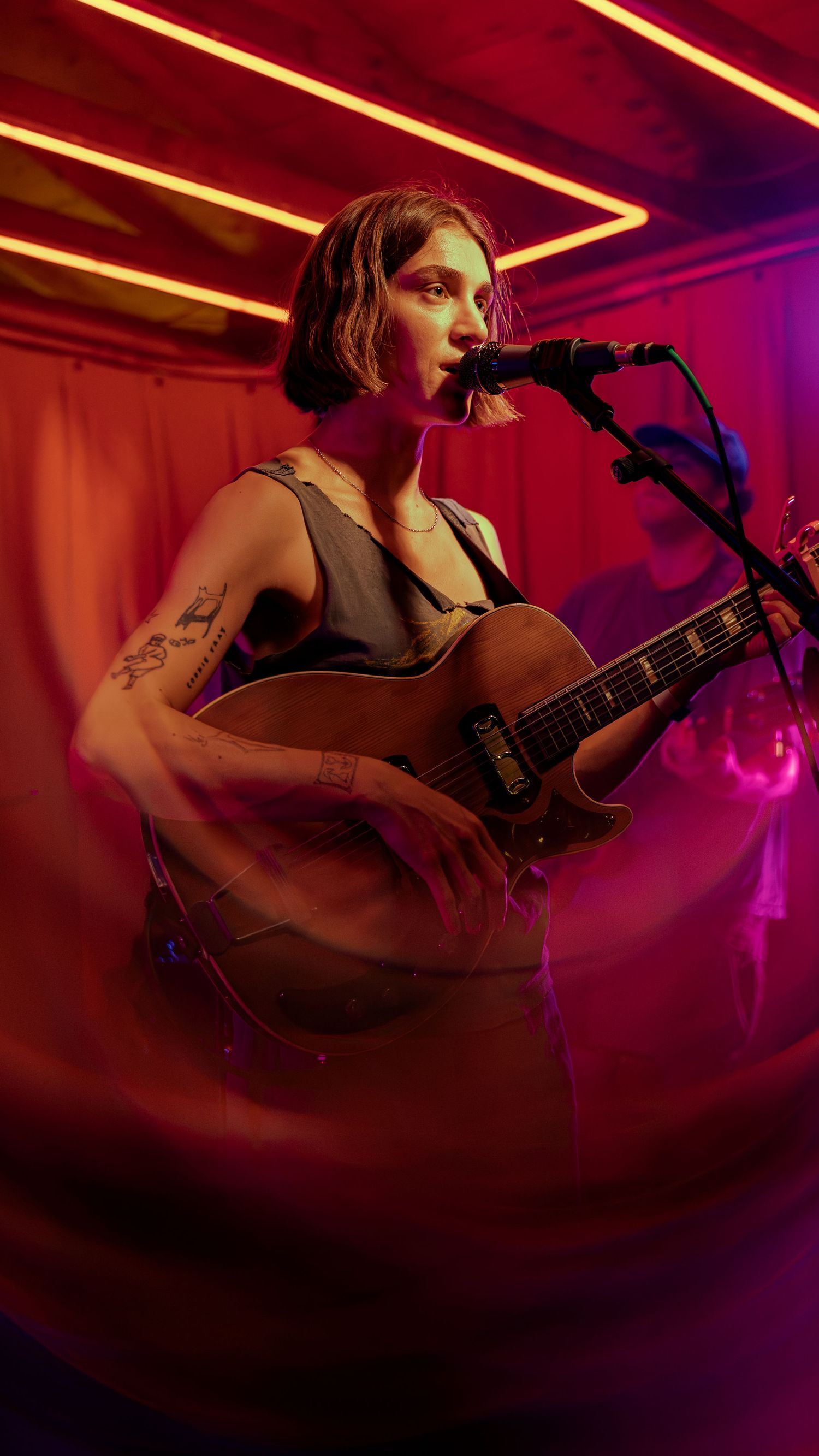 A musician performs on stage with a guitar under dramatic red and purple lighting during a live concert performance.