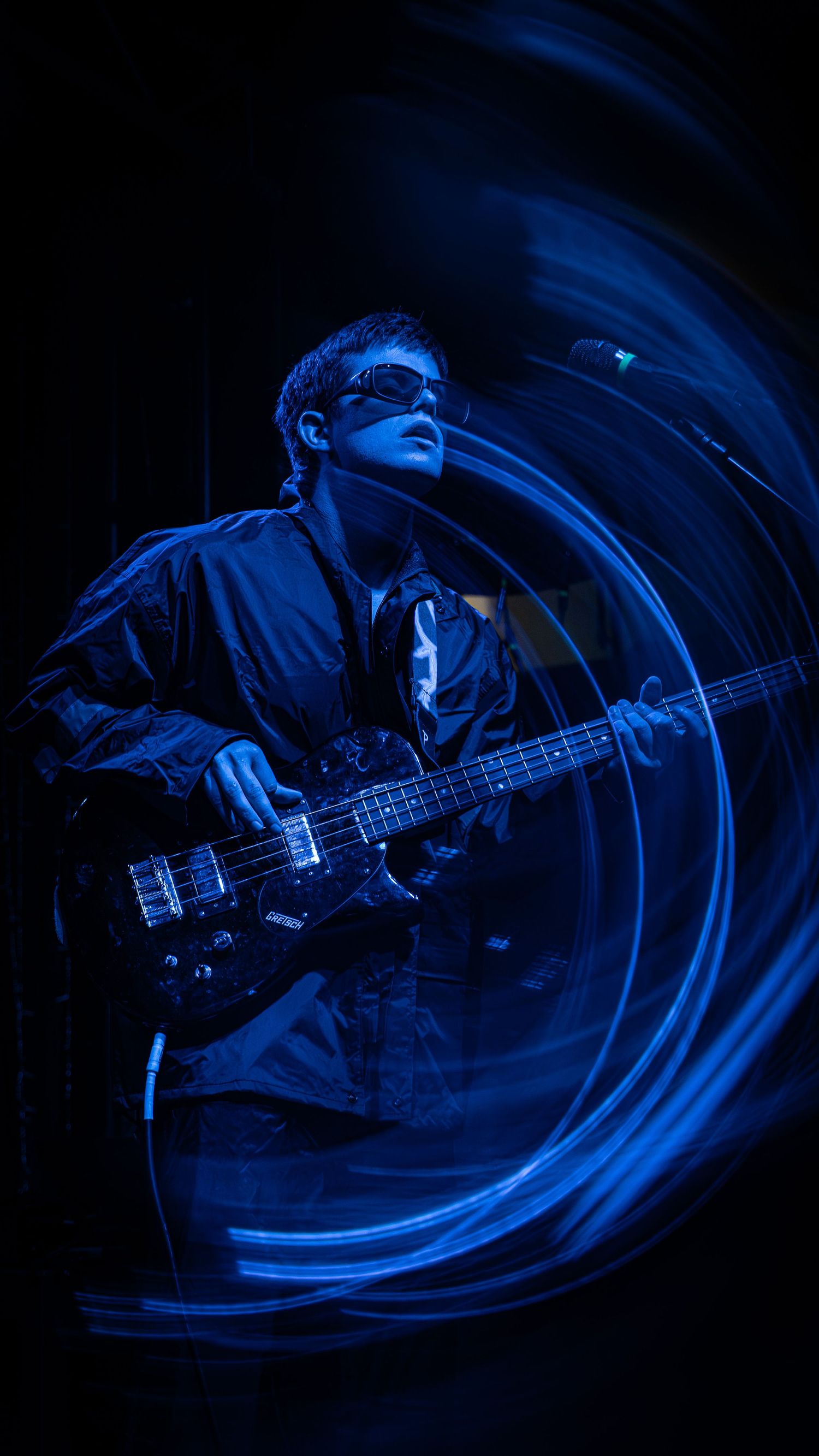 A musician plays bass guitar on stage bathed in dramatic blue lighting with swirling circular light effects in the background.