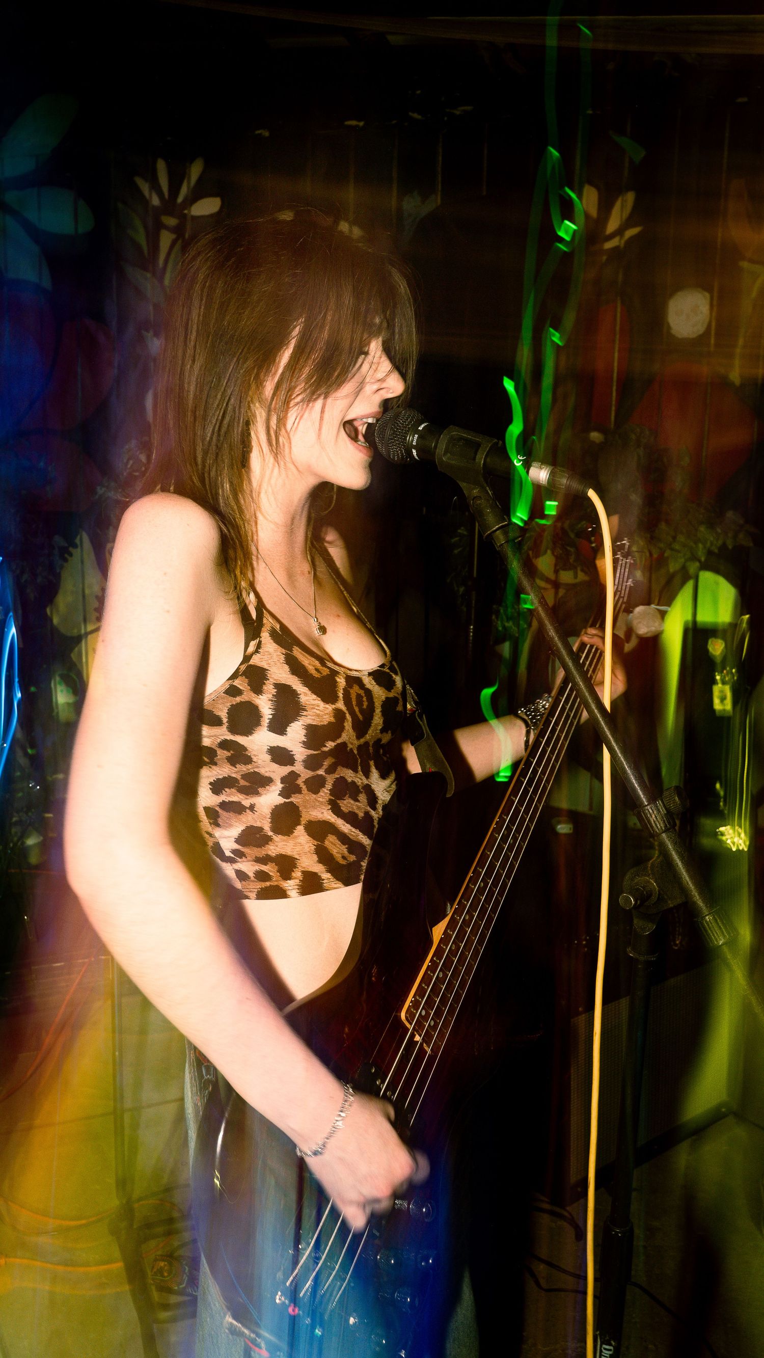 Bass player in leopard print performs on stage with green and blue light trails.