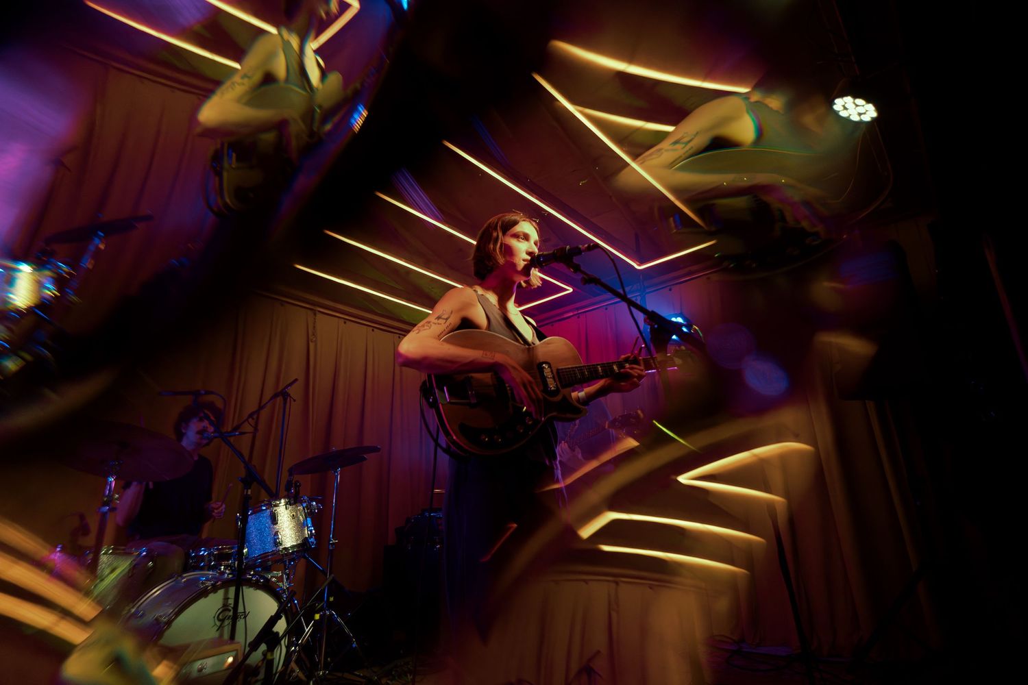 A musician performs on stage under dramatic lighting with angular wooden ceiling beams and purple spotlights creating atmosphere.