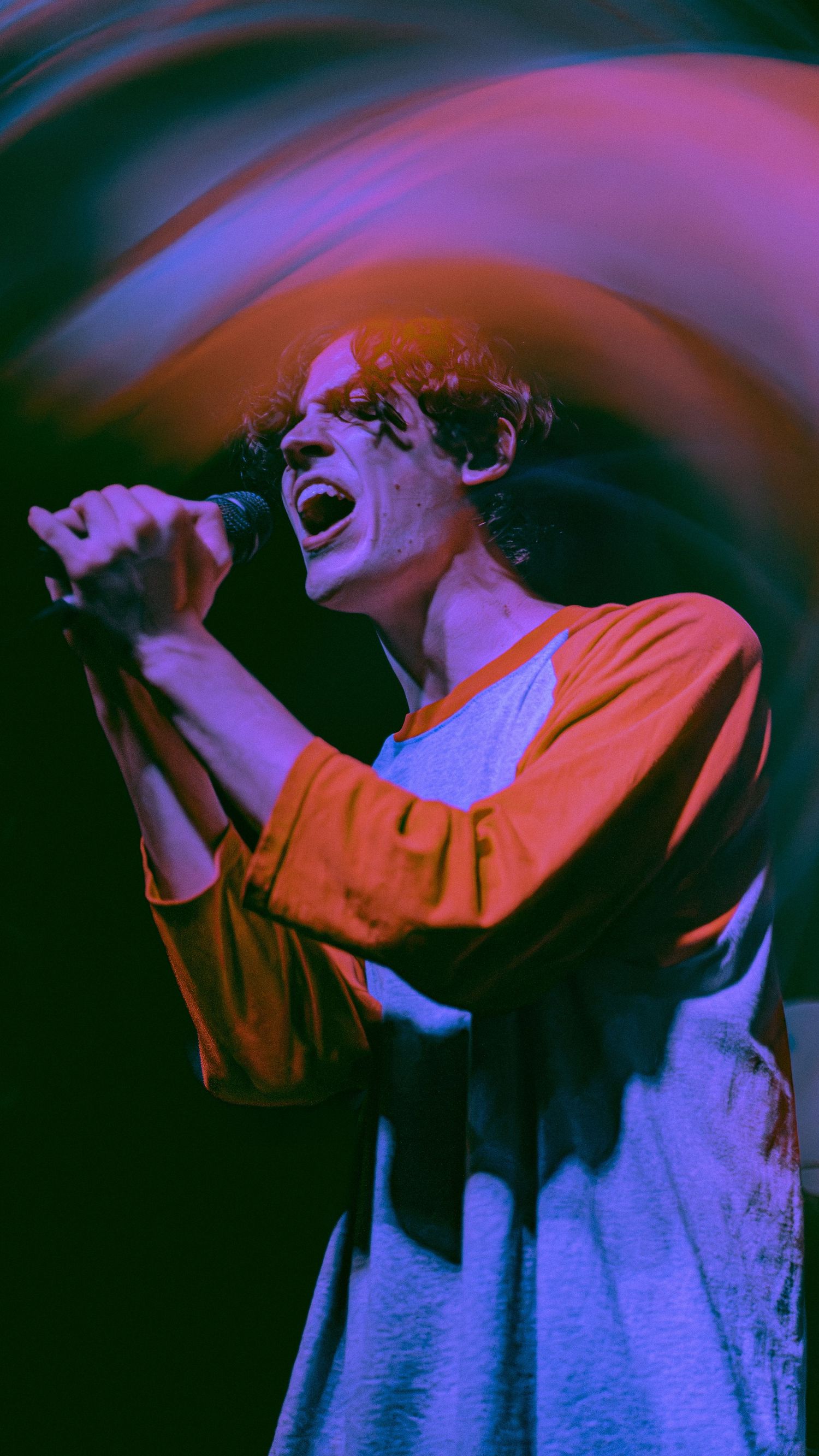 Concert performer in orange shirt singing passionately under dramatic purple stage lighting during live performance.