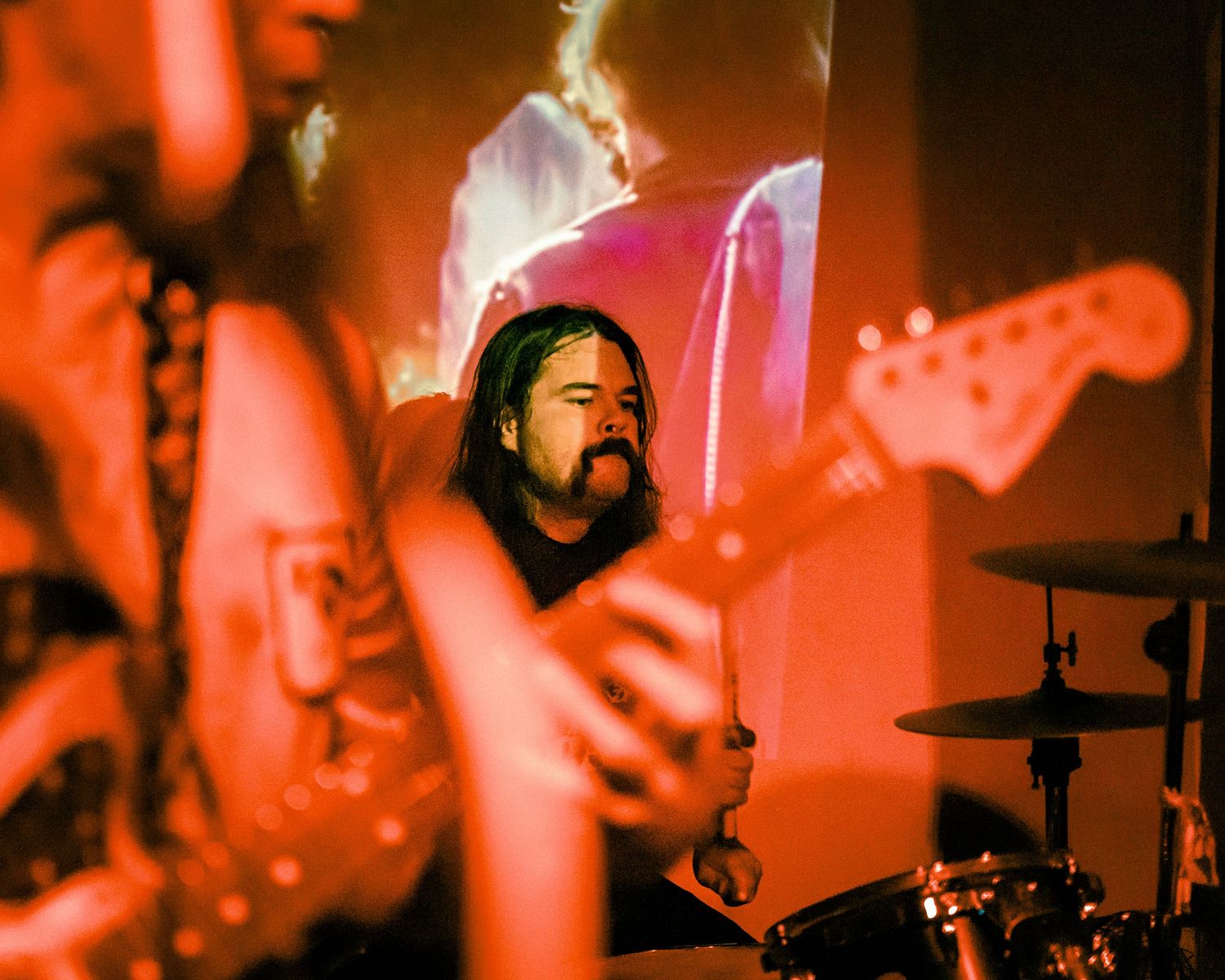 Musician performing on stage bathed in intense red lighting with guitar silhouette visible.