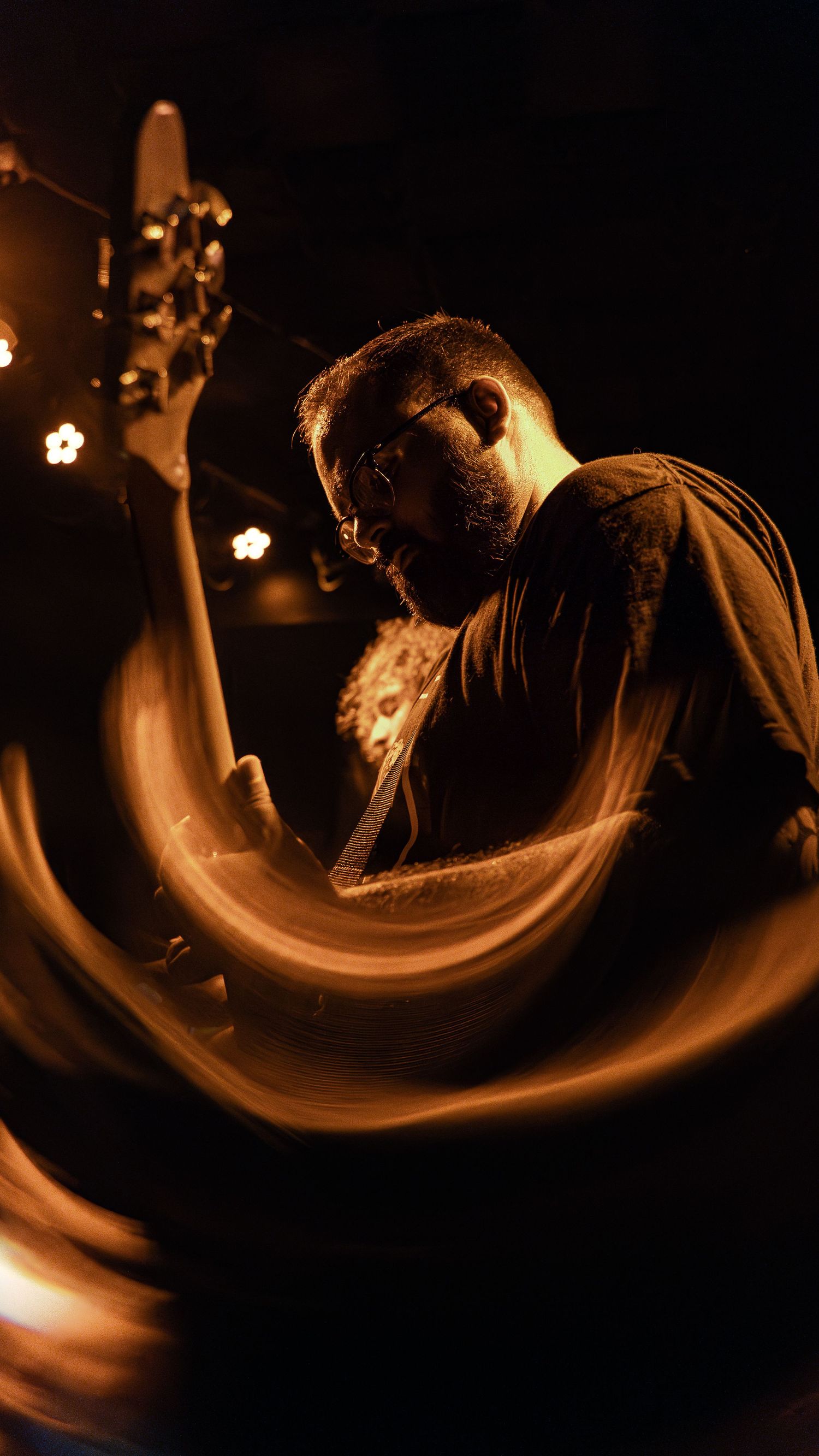 Musician playing bass guitar in dramatic lighting with swirling orange light trails creating an artistic motion effect.