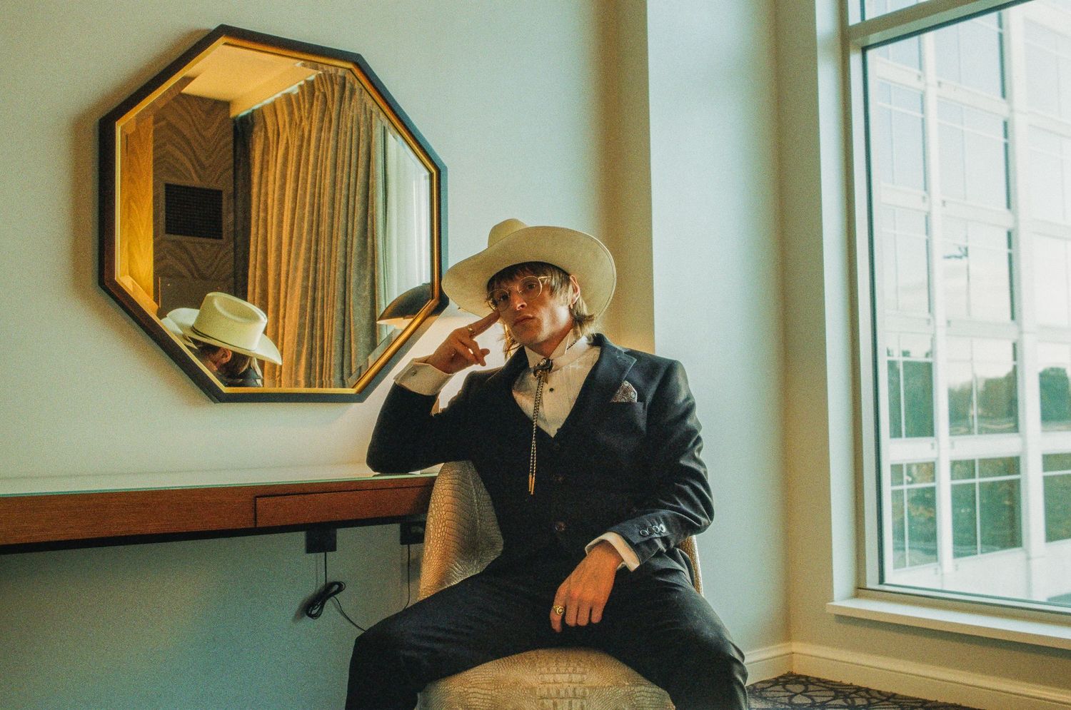 Person in a black blazer and cowboy hat poses thoughtfully by a window with an octagonal mirror on the wall.