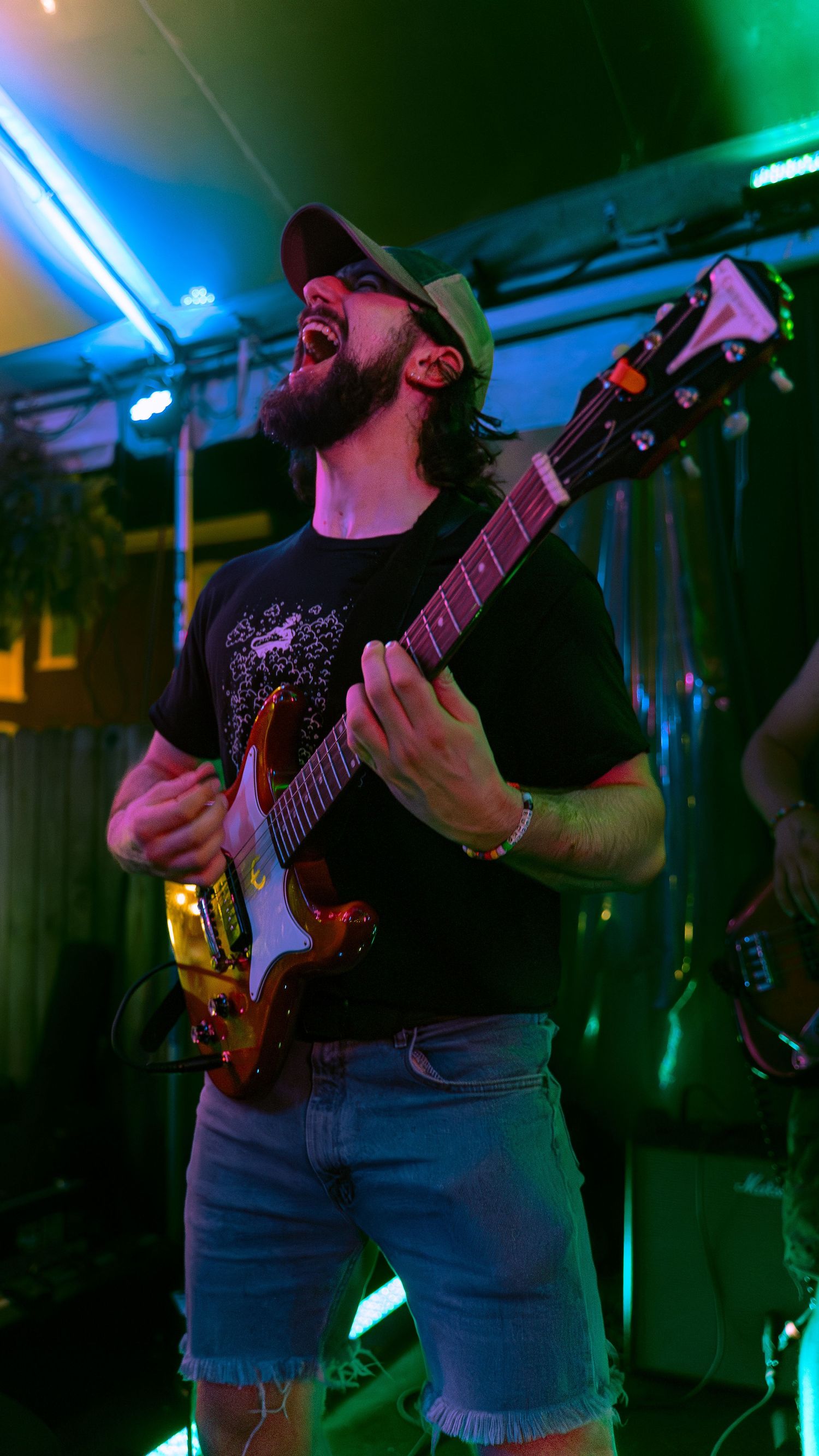 Person playing electric guitar on stage in dark lighting with blue and green ambient lighting in the background.