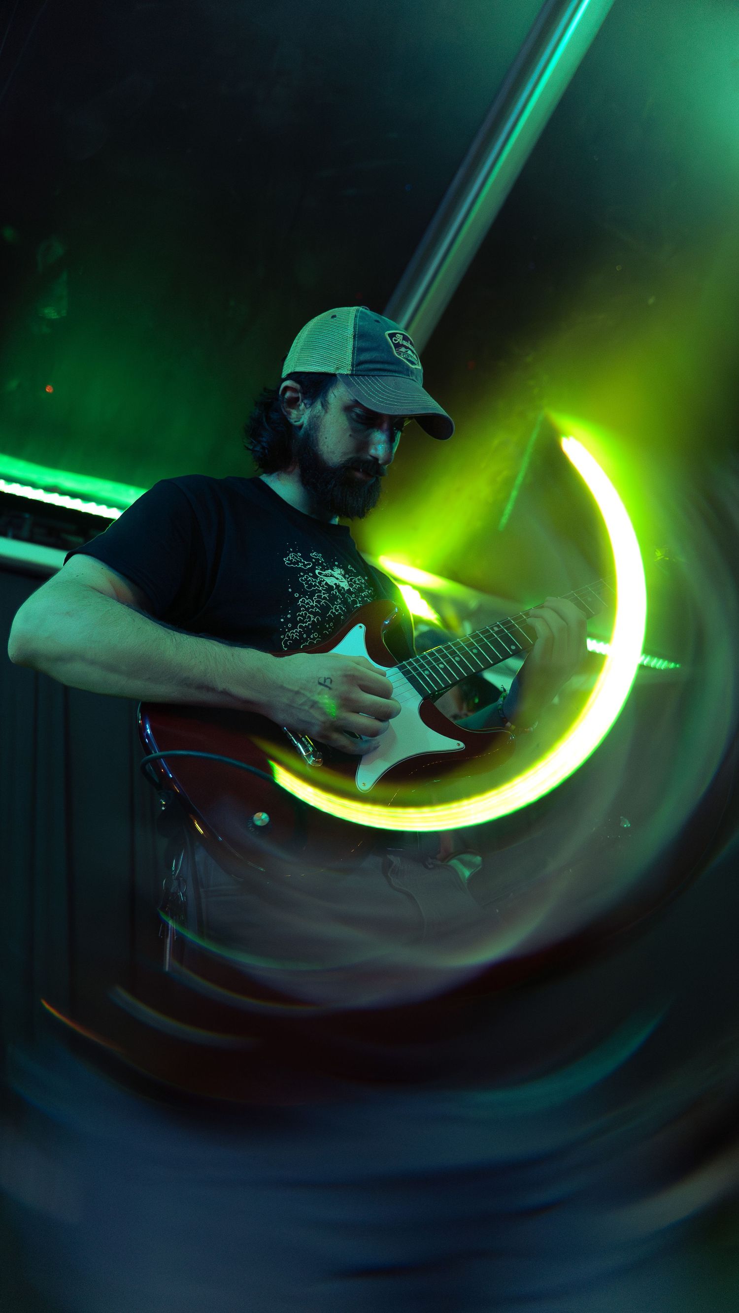Musician playing electric guitar on dark stage with dramatic green lighting creating glowing curved light trails.