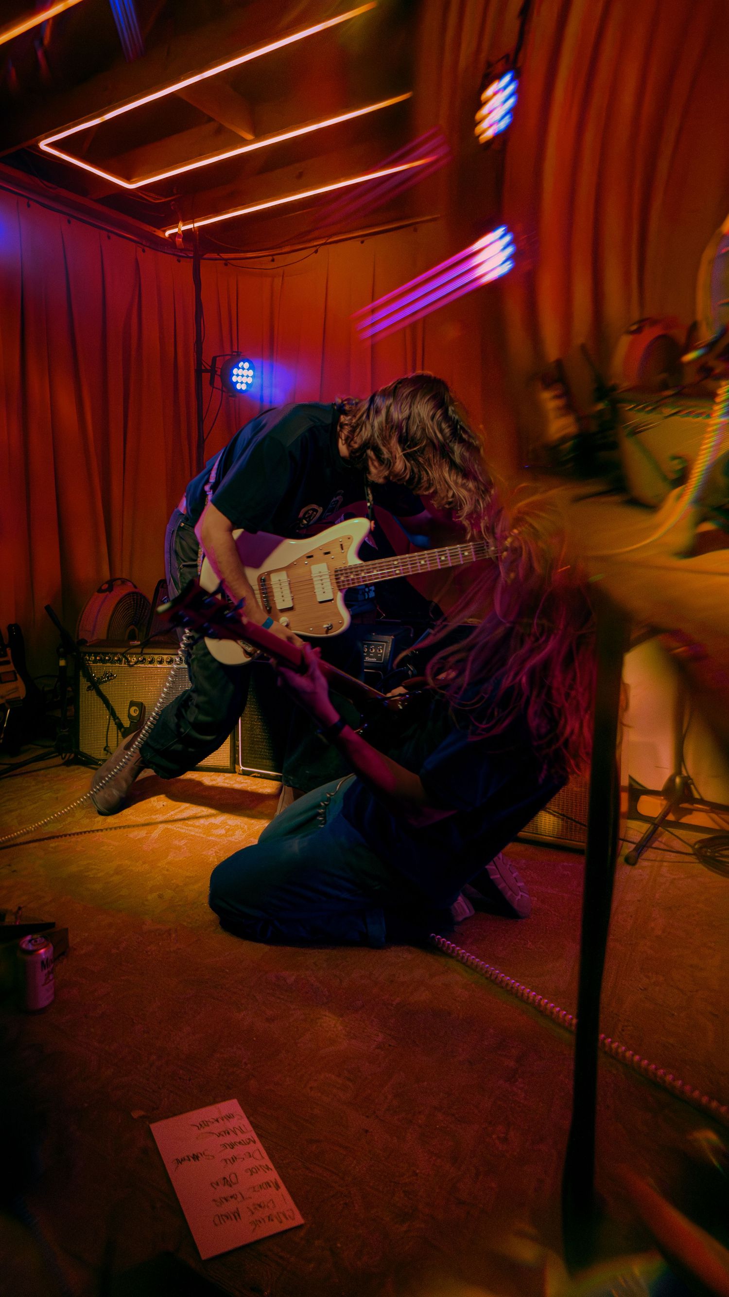 A musician performs energetically with an electric guitar in a dimly lit venue with red and purple lighting.