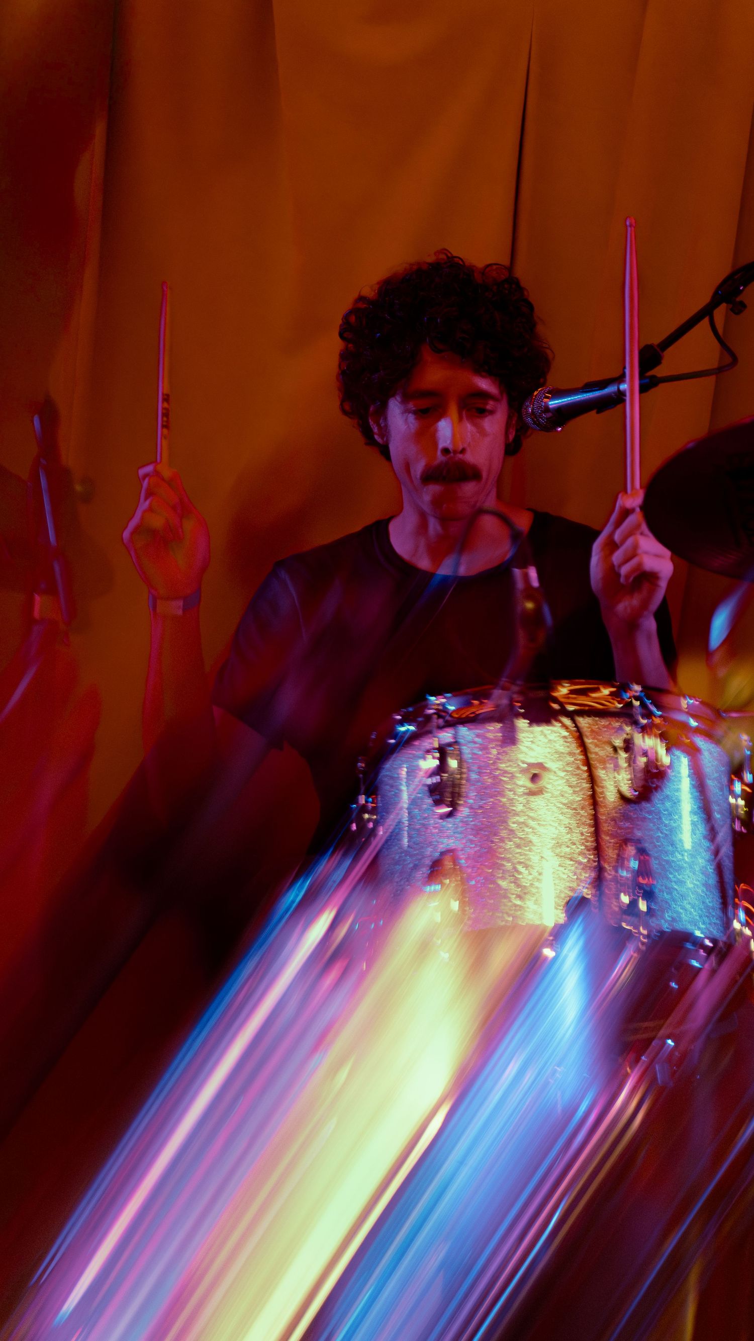 A silhouetted drummer performing behind colorful neon light streaks in blue, yellow and pink during a live show.