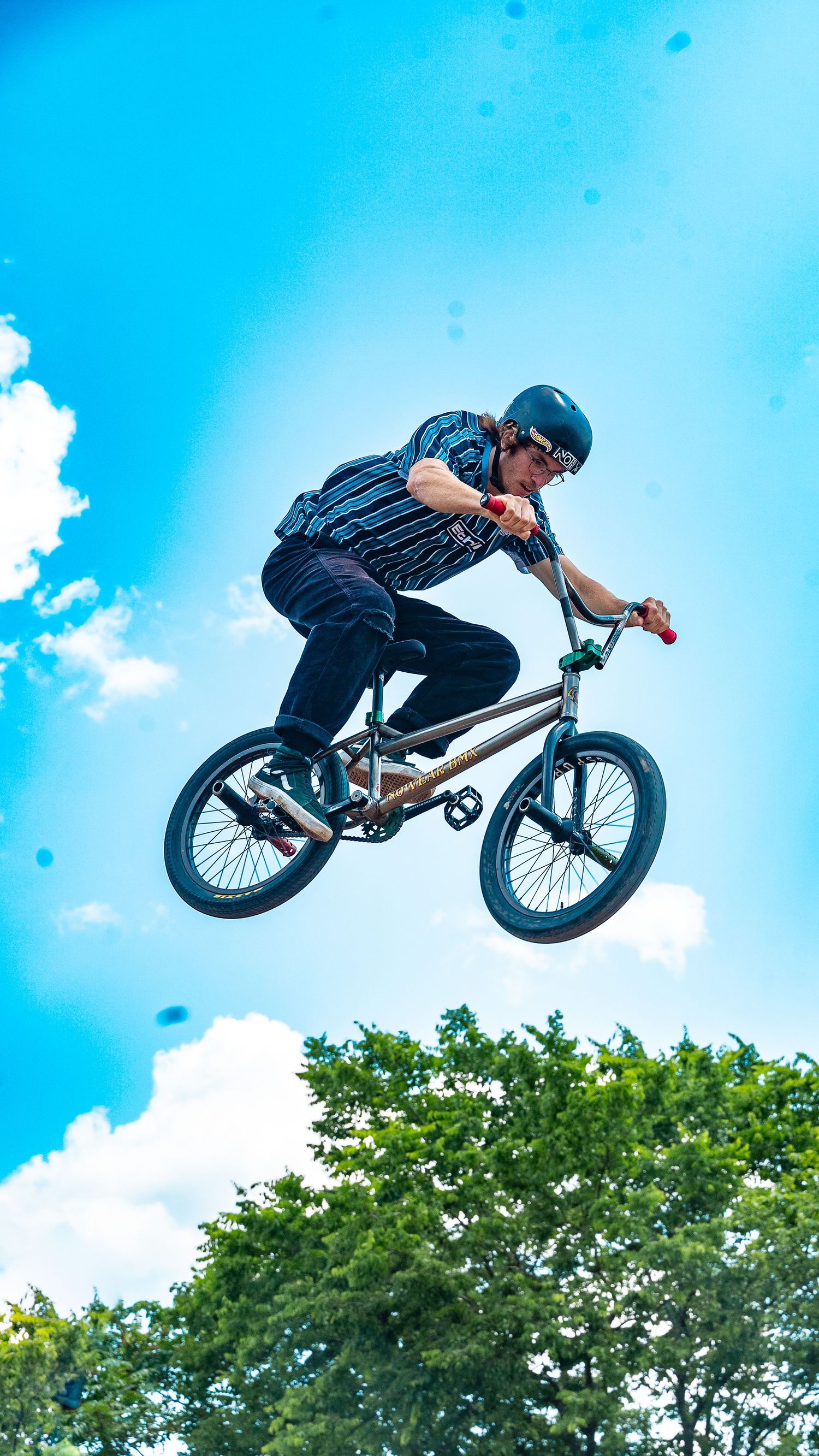 BMX biker executes high-flying jump trick against summer sky and trees.
