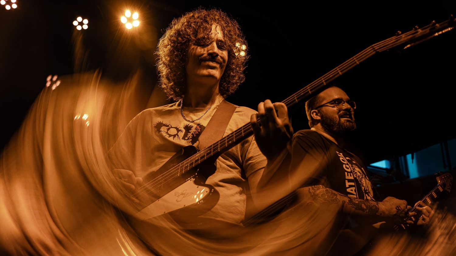 Musicians perform energetically on stage with electric guitars under dramatic lighting creating motion blur effects.