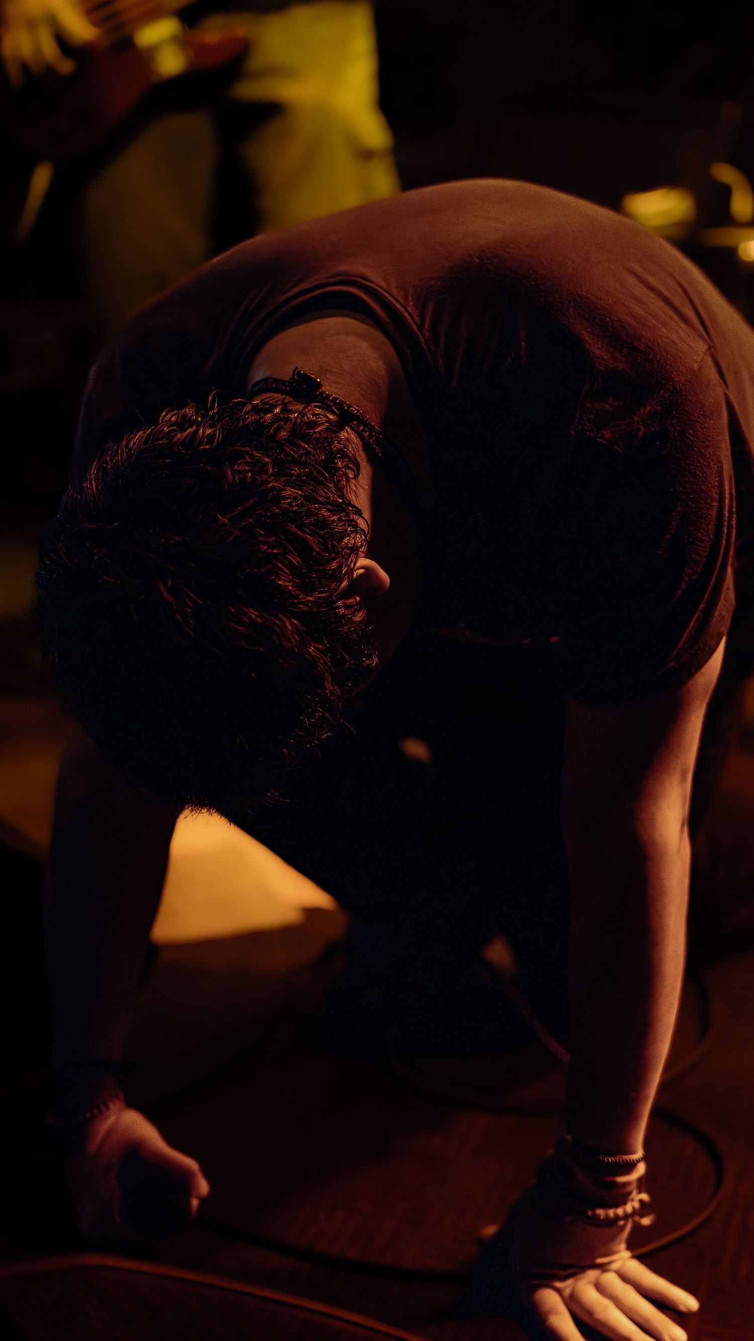 A darkly lit figure performs a yoga or stretching pose on a mat in atmospheric low lighting.