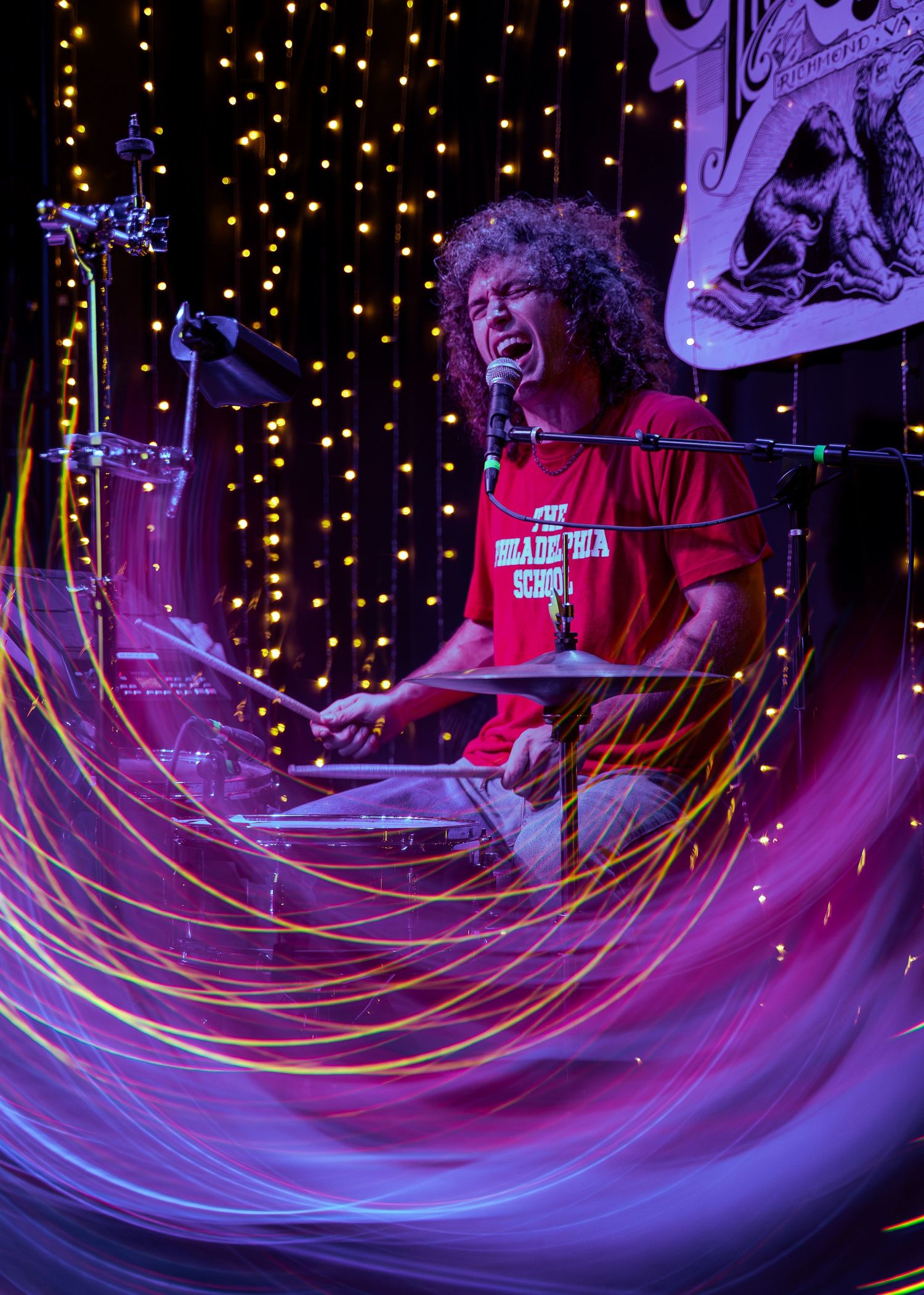 A drummer performs energetically in purple and red stage lighting with twinkling lights in the background.
