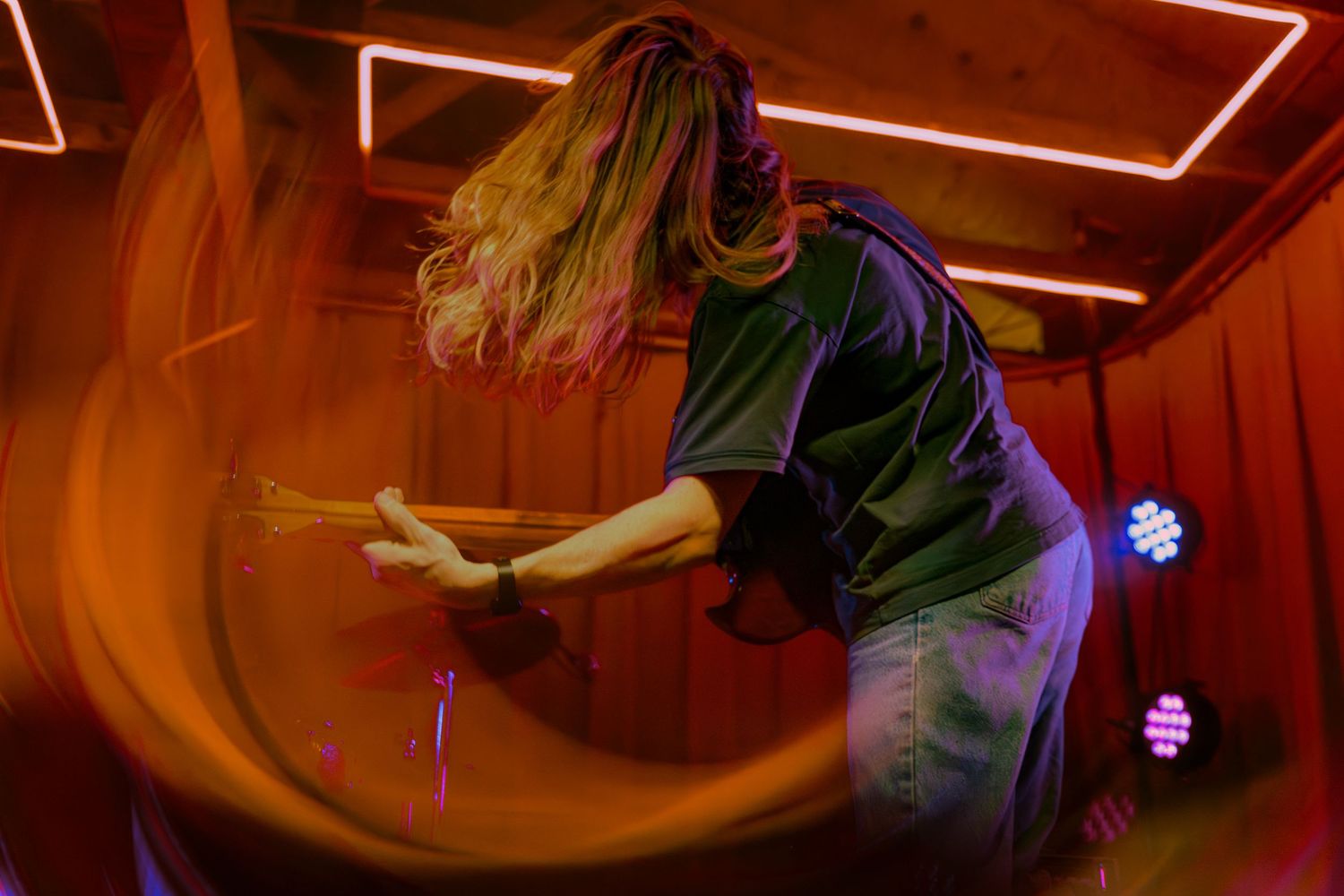 A performer with flowing hair spins dynamically on stage under red lighting at a nightclub.