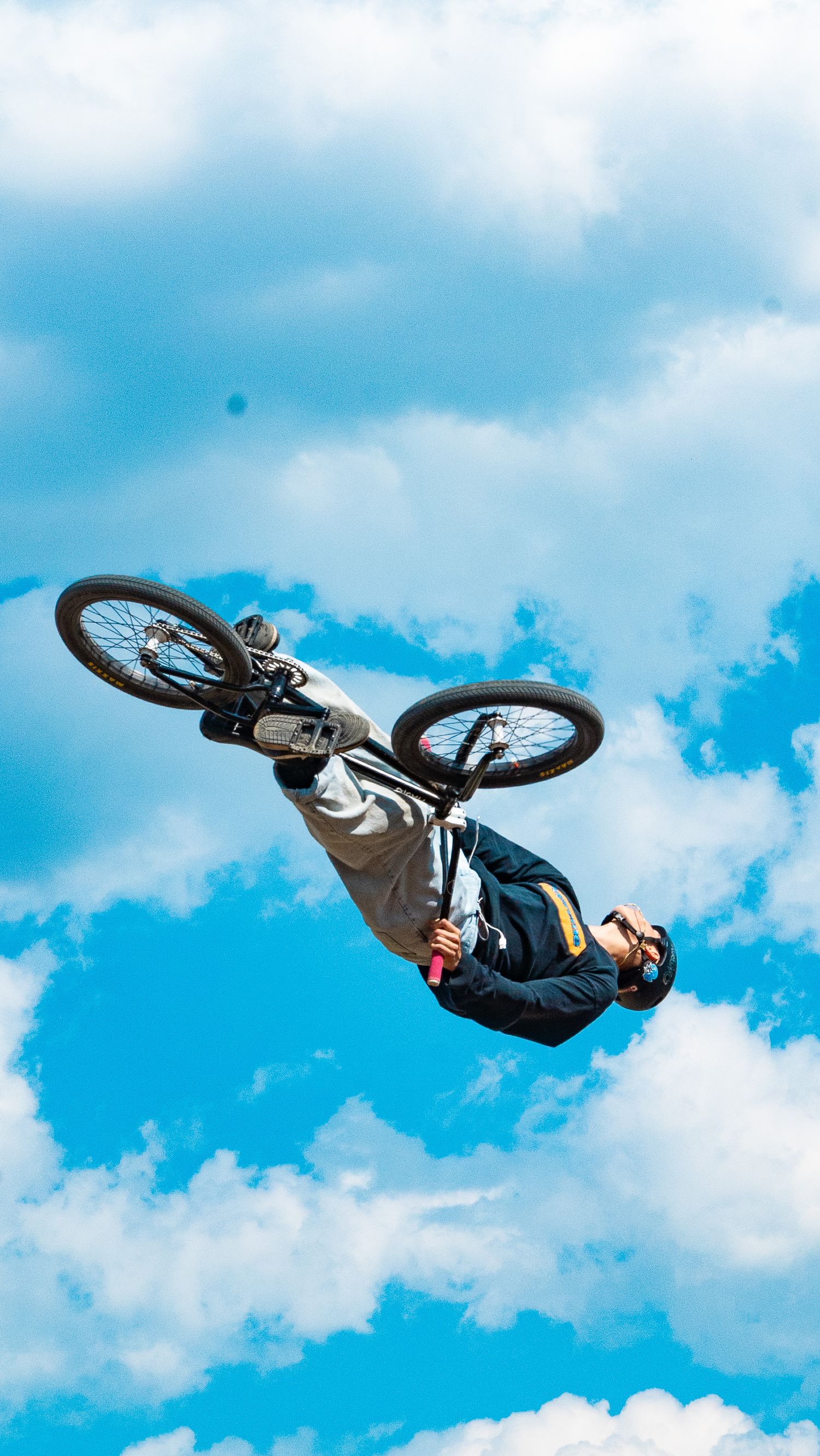 BMX rider rotates bike sideways in mid-air against bright blue sky with white clouds.