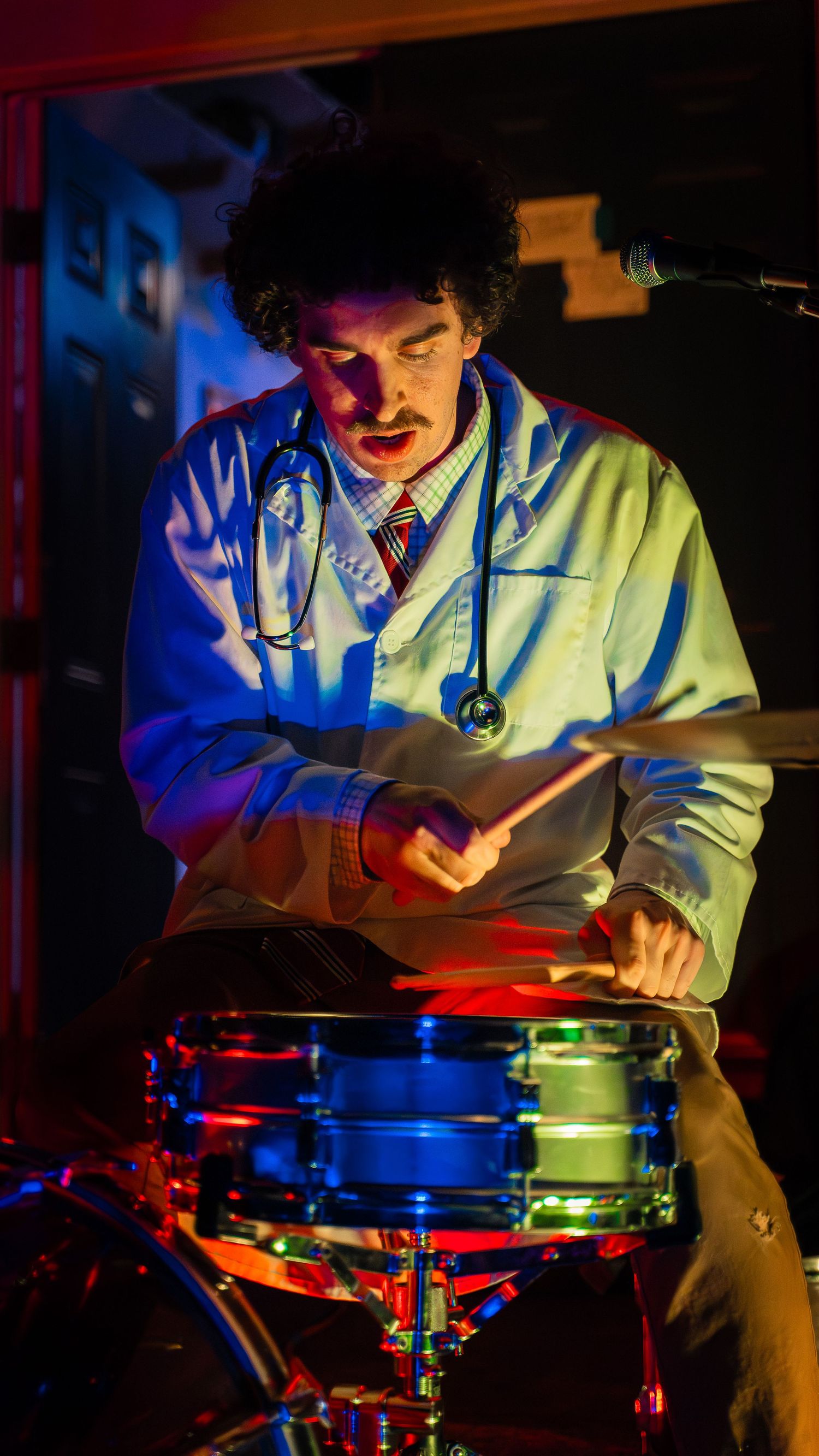 Drummer performing behind a drum kit illuminated by dramatic blue and green stage lighting.