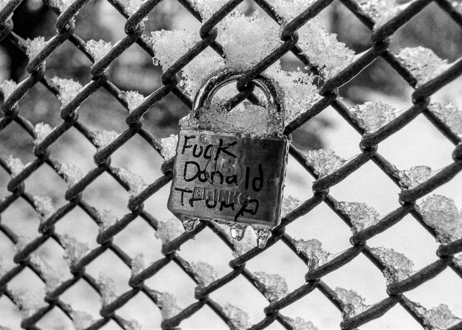 A rusty padlock with graffiti text engraved on it hangs from a chain link fence in black and white.
