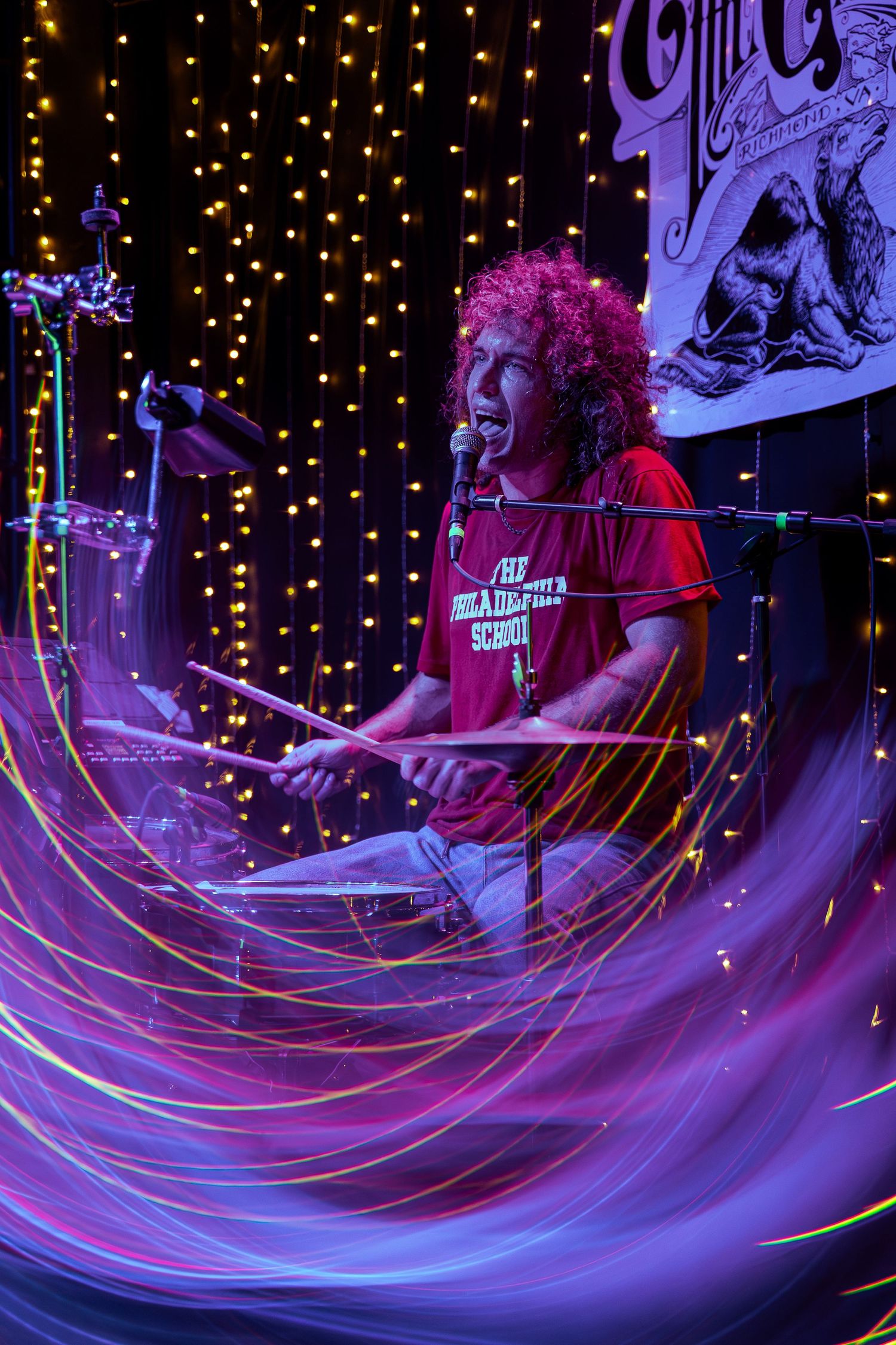 A drummer performs energetically on stage with twisting purple light trails and glowing string lights in the background.