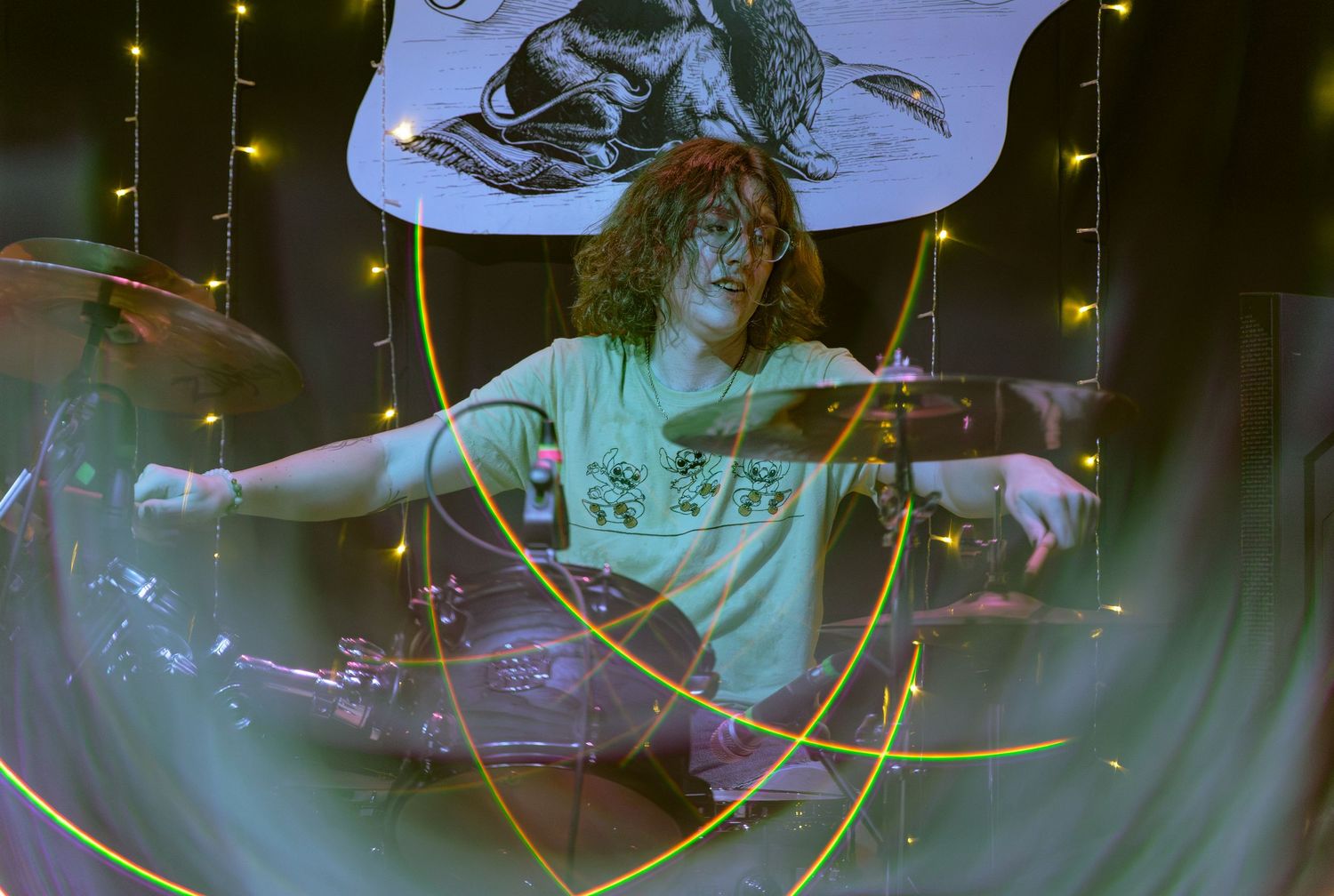A drummer performs energetically behind a drum kit with colorful stage lighting and motion blur effects.