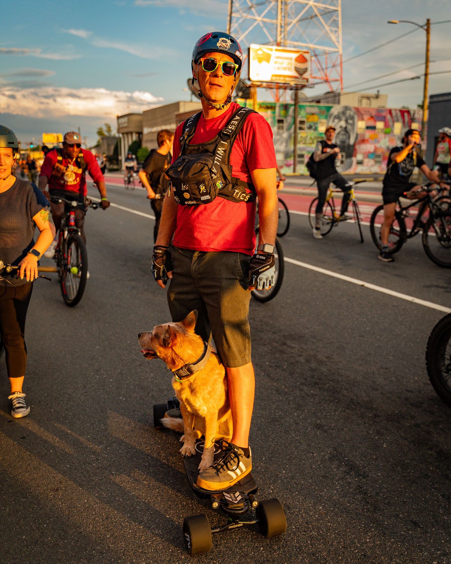 Skateboarder with dog stands on street during group bike ride at sunset.