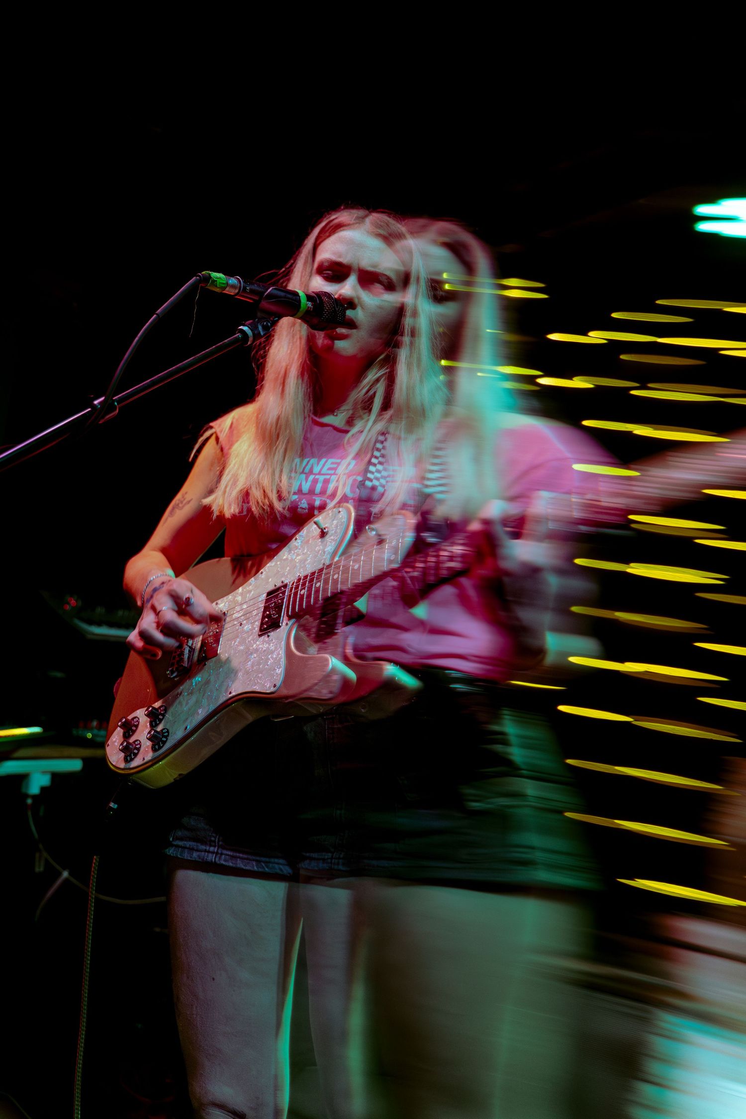 Blurred motion shot of musician playing an electric guitar with vibrant yellow and pink lighting effects on stage.